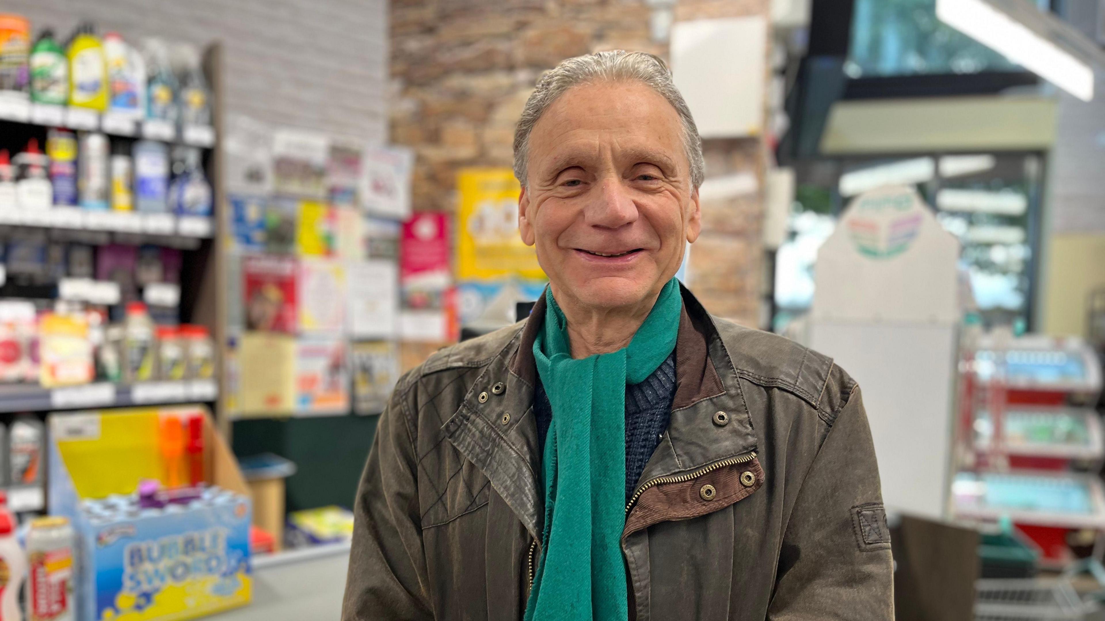 A man wearing a green scarf and brown leather jacket stands in a petrol station shop.