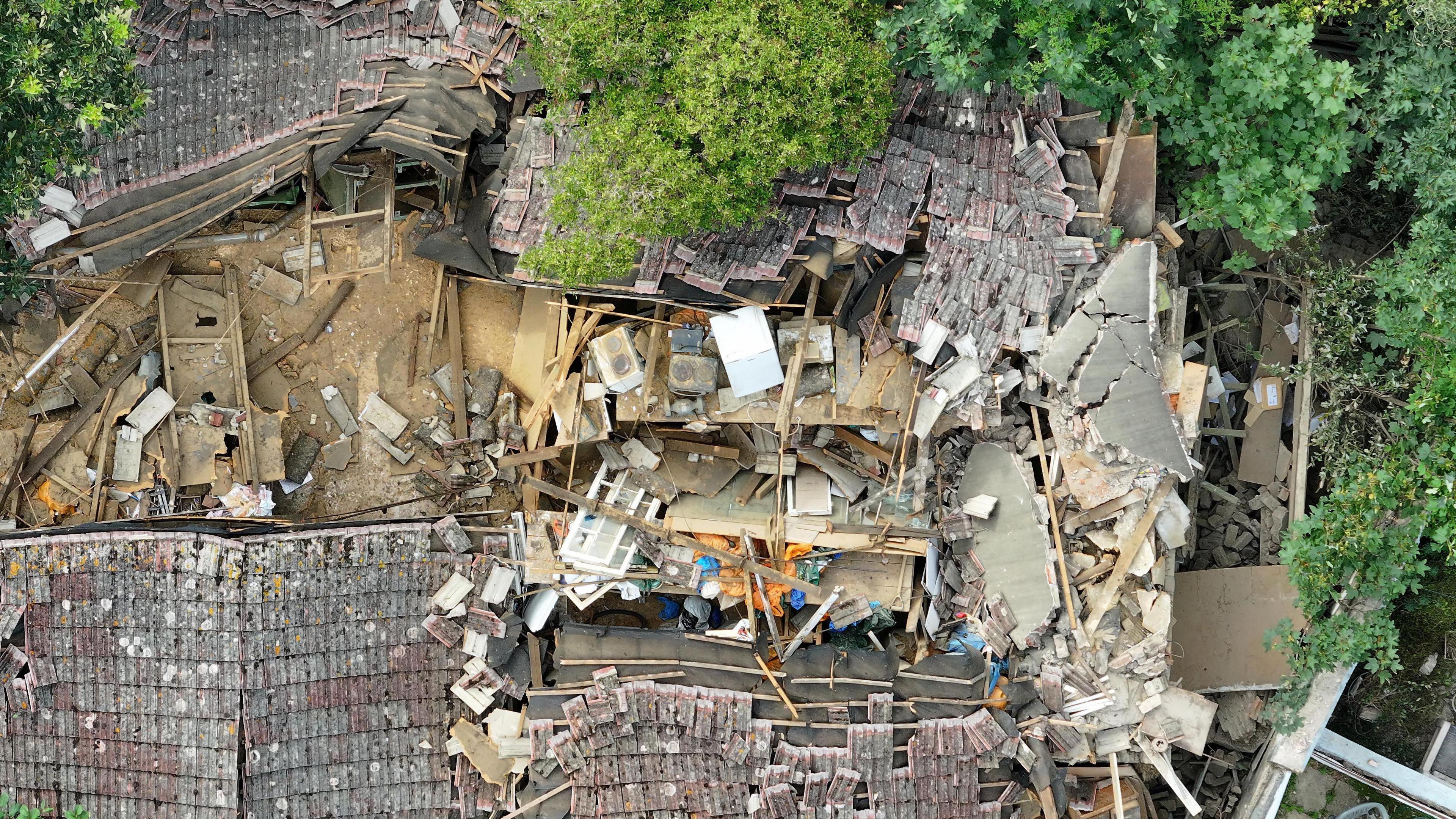 The partially collapsed building is viewed from an aerial perspective, showing an exposed first floor. Dust and debris are scattered everywhere, with wooden beams, walls, slate and stone visibly poking out of collapsed mounds. Leafy trees and shrubbery surround the building's perimeter, with fallen debris resting against nearby fences to neighbouring properties.
