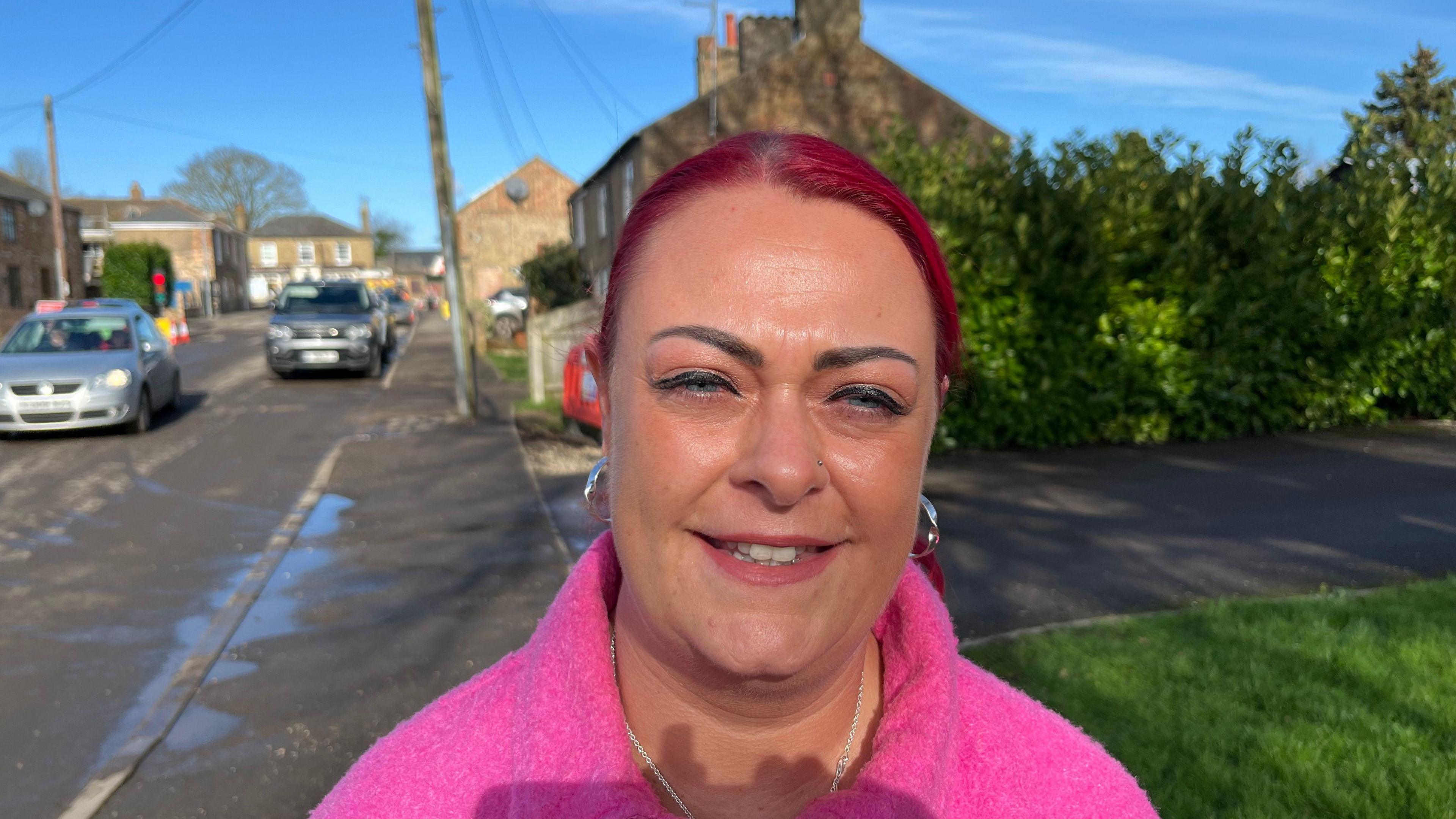 Hannah Cooke, 41 has dyed red hair pulled back tightly over her head she has bold black eyebrows and has dangling silver earrings. She is wearing a fleecy pink top and has a silver chain around her neck. Behind her is a roadway with cars parked and one silver car driving past her there are houses visible too.