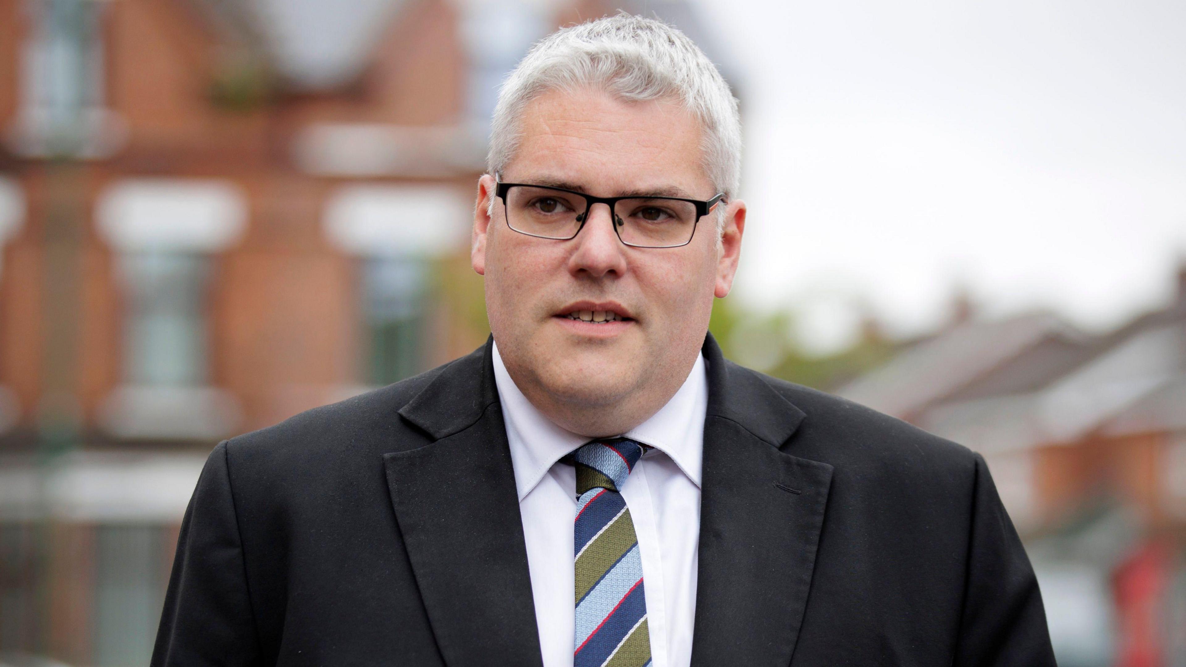 Gavin Robinson wearing a suit and tie looking slightly off camera with a neutral expression. Some buildings in the background are out of focus.