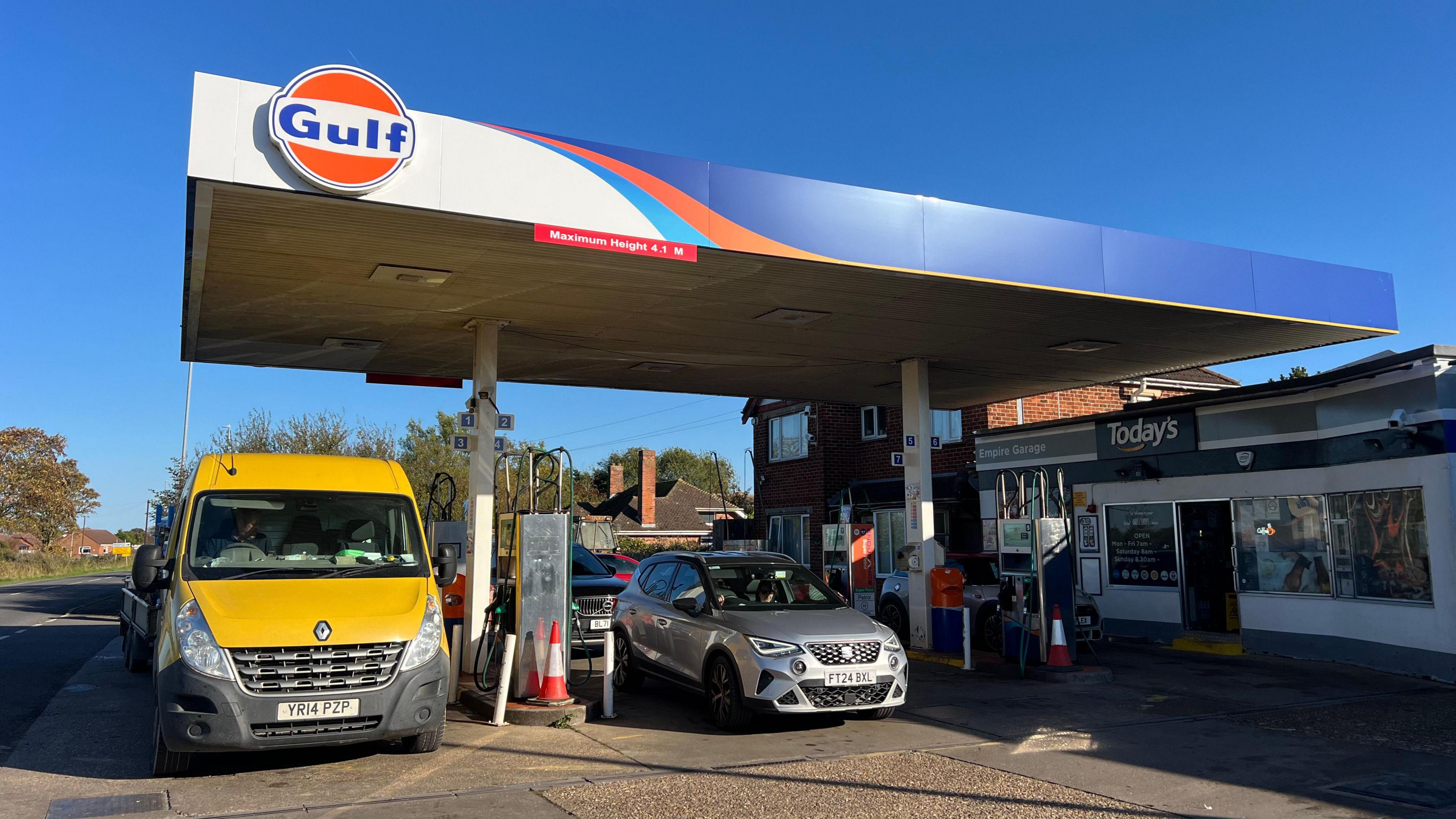 A Gulf petrol station. A yellow van is parked on the left of the photo and a silver car is parked on the right of it. There is a 'today's' shop on the right of the photo. The sky is blue.