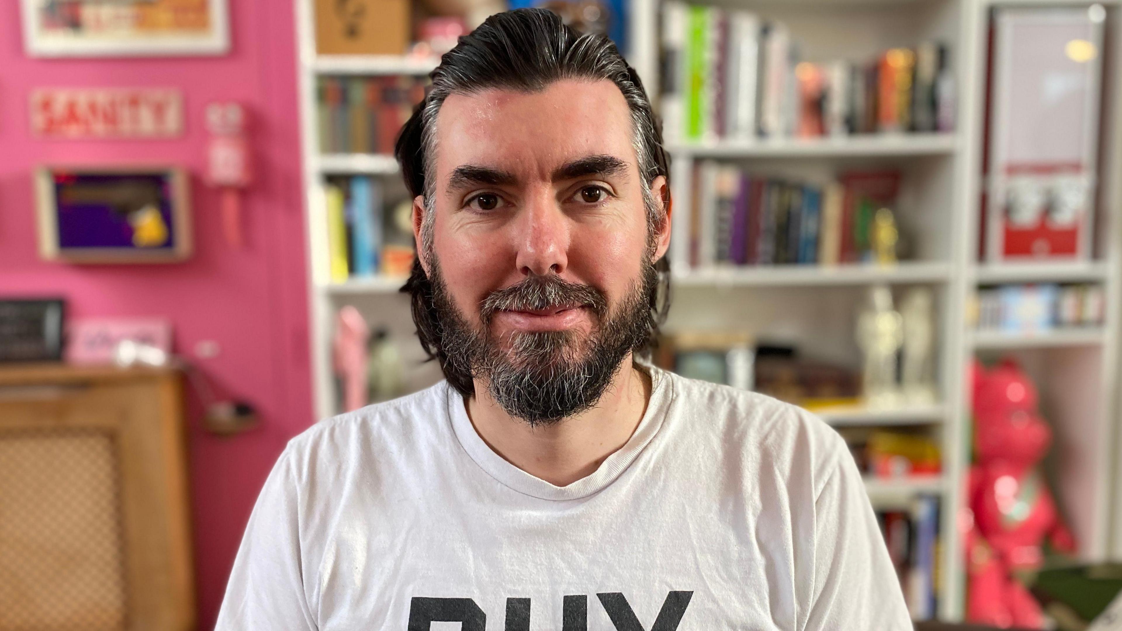 Adam Carthy has brown hair and beard. He is wearing a white T-shirt that says BHX and is sitting in front of a bookshelf.