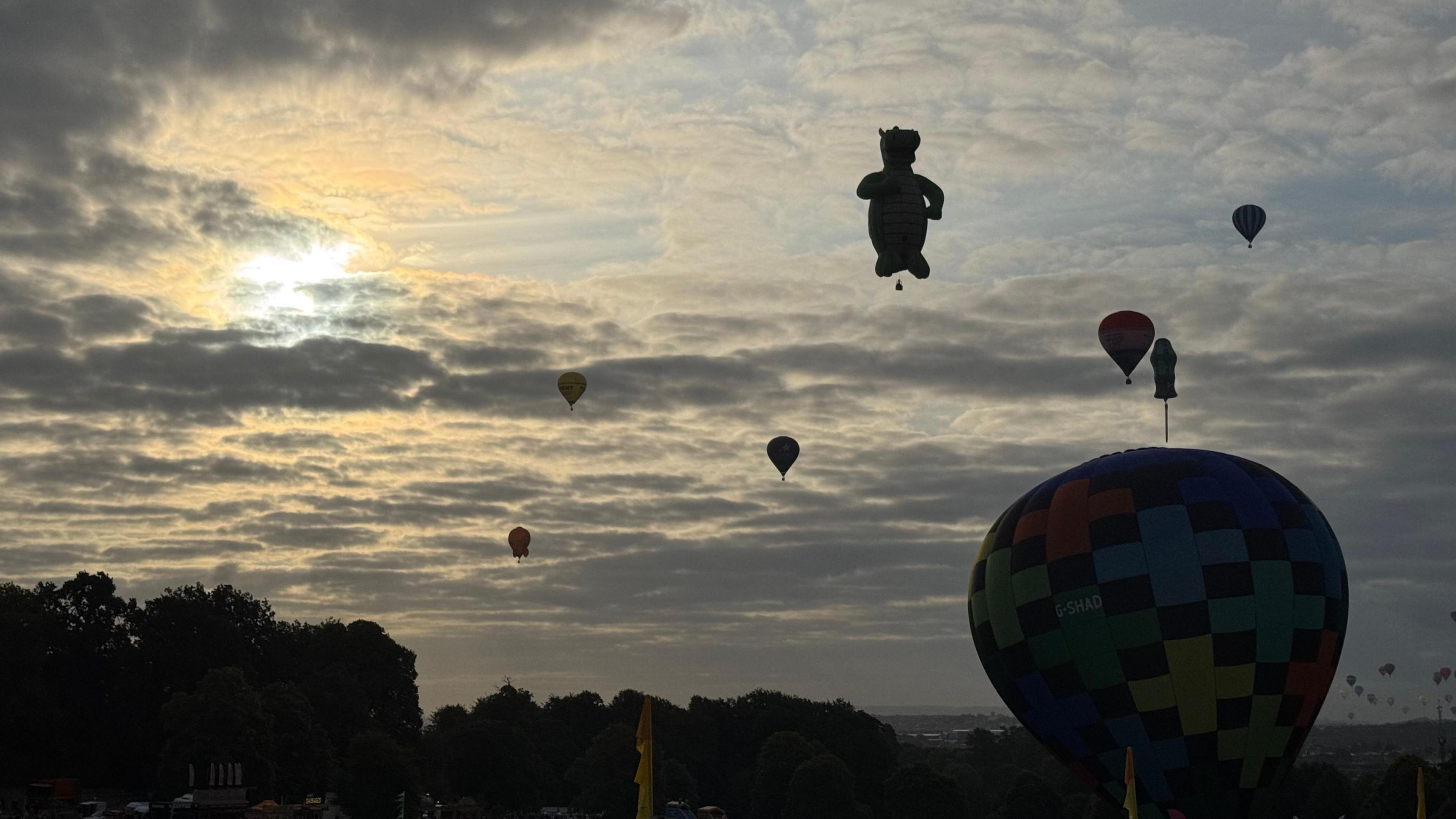 Eight hot air balloons, with one in the shape of a dragon and another as a screwdriver, silhouetted against the sky on a cloudy day.