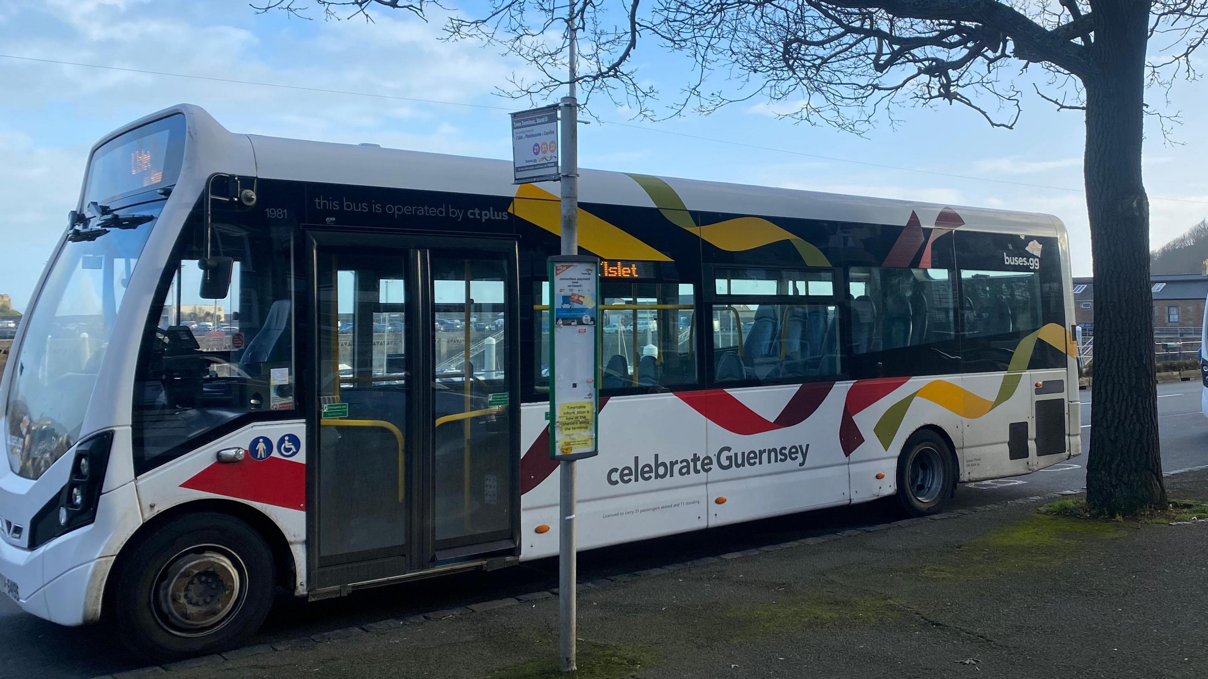 An empty bus is parked at a bus stop. Lettering on the bus reads 'celebrate Guernsey'. 