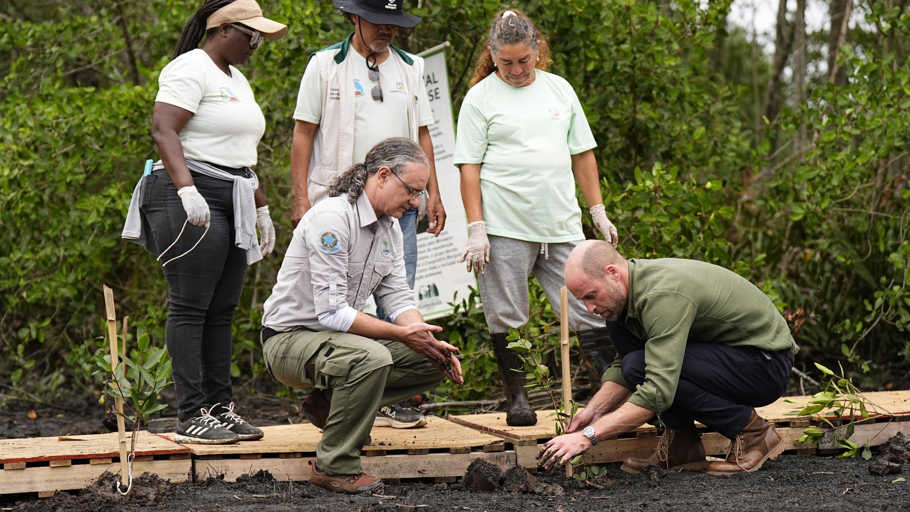 William crouched on the right, facing left and he plants something in the soil. Crouched opposite him is a man with long grey hair tied up and glasses. Behind them, is a woman wearing a visor and glasses, a man in a hat, and a woman. They are outdoors. In the background are multiple shrubs.