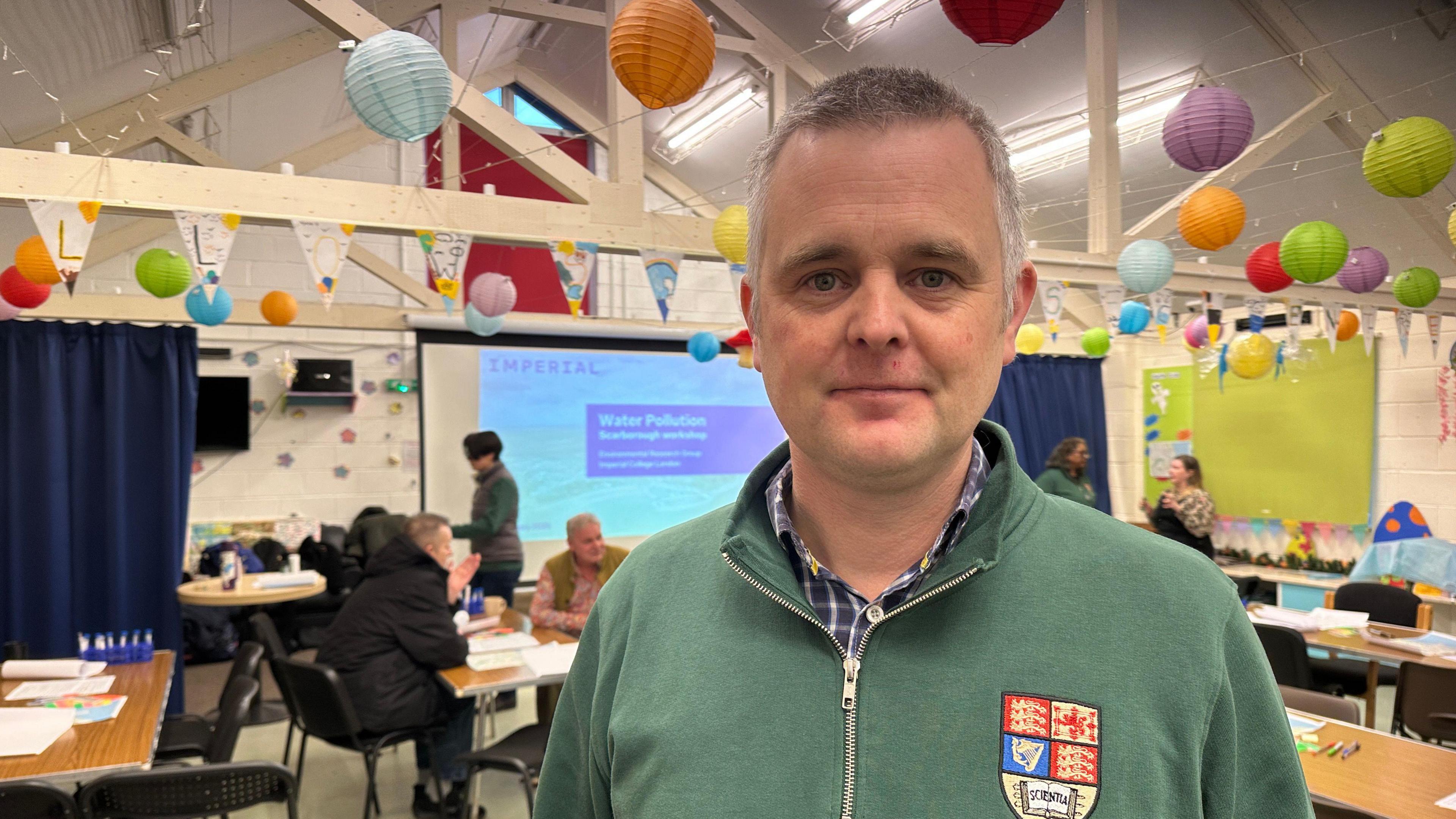 Man in a green fleece stands in a classroom and smiles at the camera.