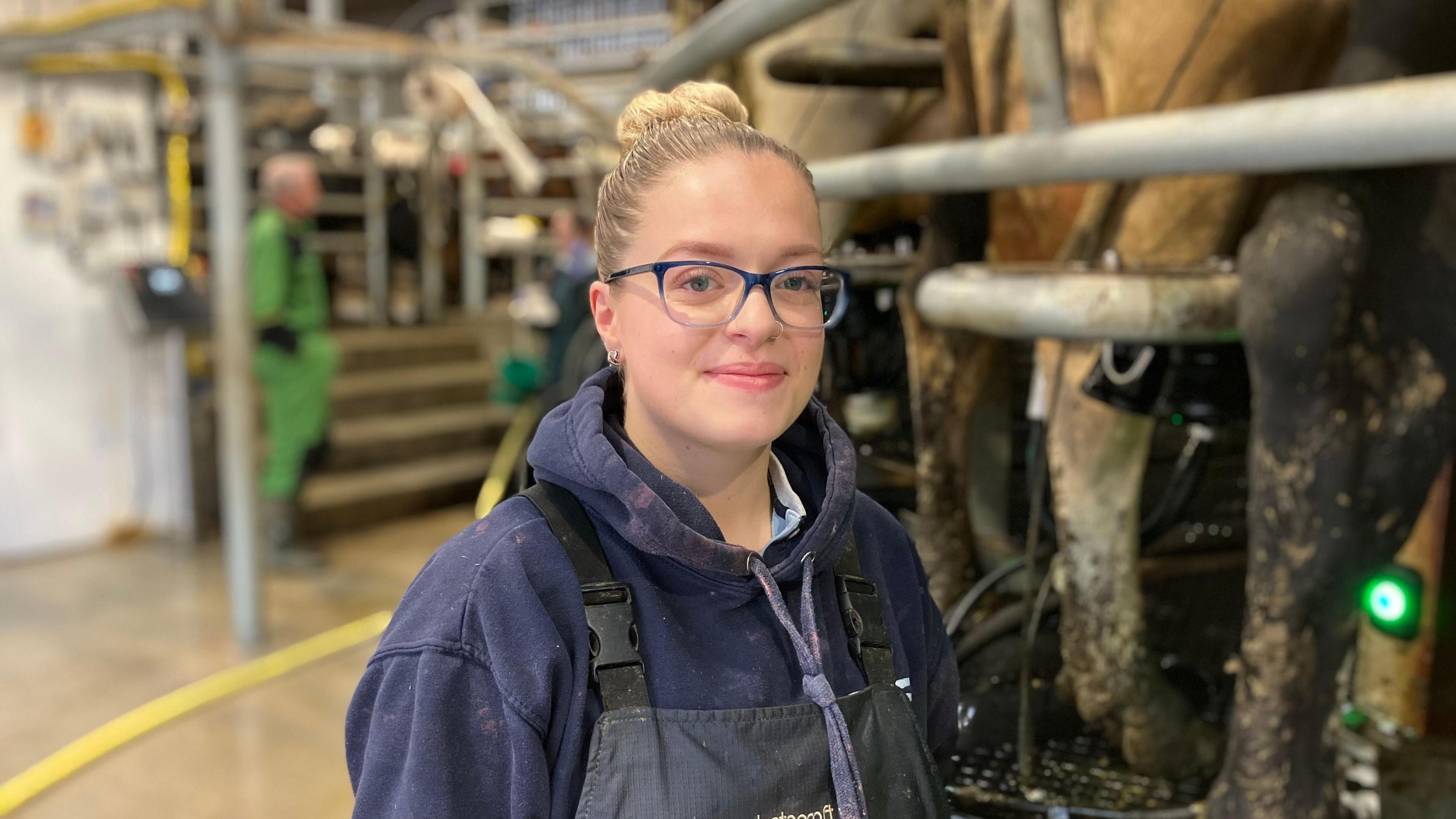 A woman stands in a milking parlour on a farm, there are cows on a raised level behind her. There is a farmer in overalls in the background. She wears blue glasses, a blue hooded jumper and overalls. Her blonde hair is tied in a bun.