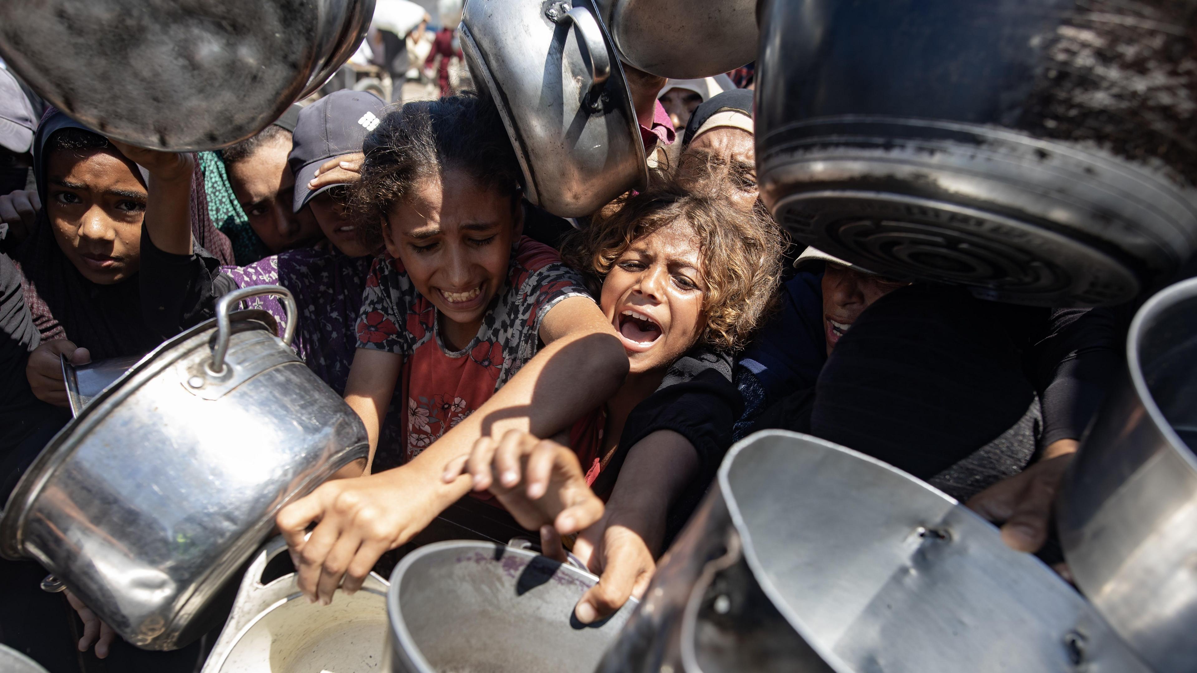 Palestinian people are tightly packed together, holding out big silver pots towards the camera.