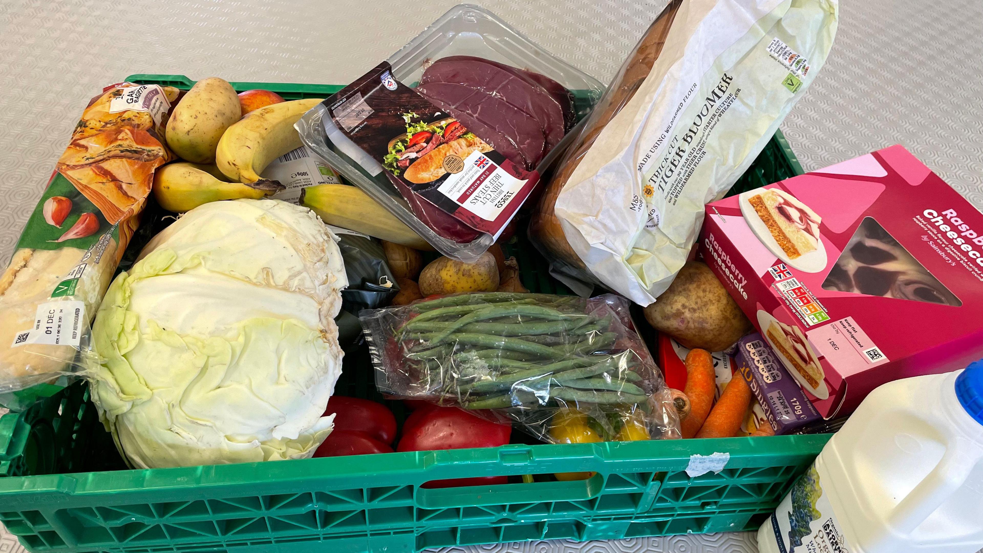 A green pallet containing cabbage, milk, bread, steak, bananas, potatoes, garlic bread, cheesecake, green beans, carrots and peppers.