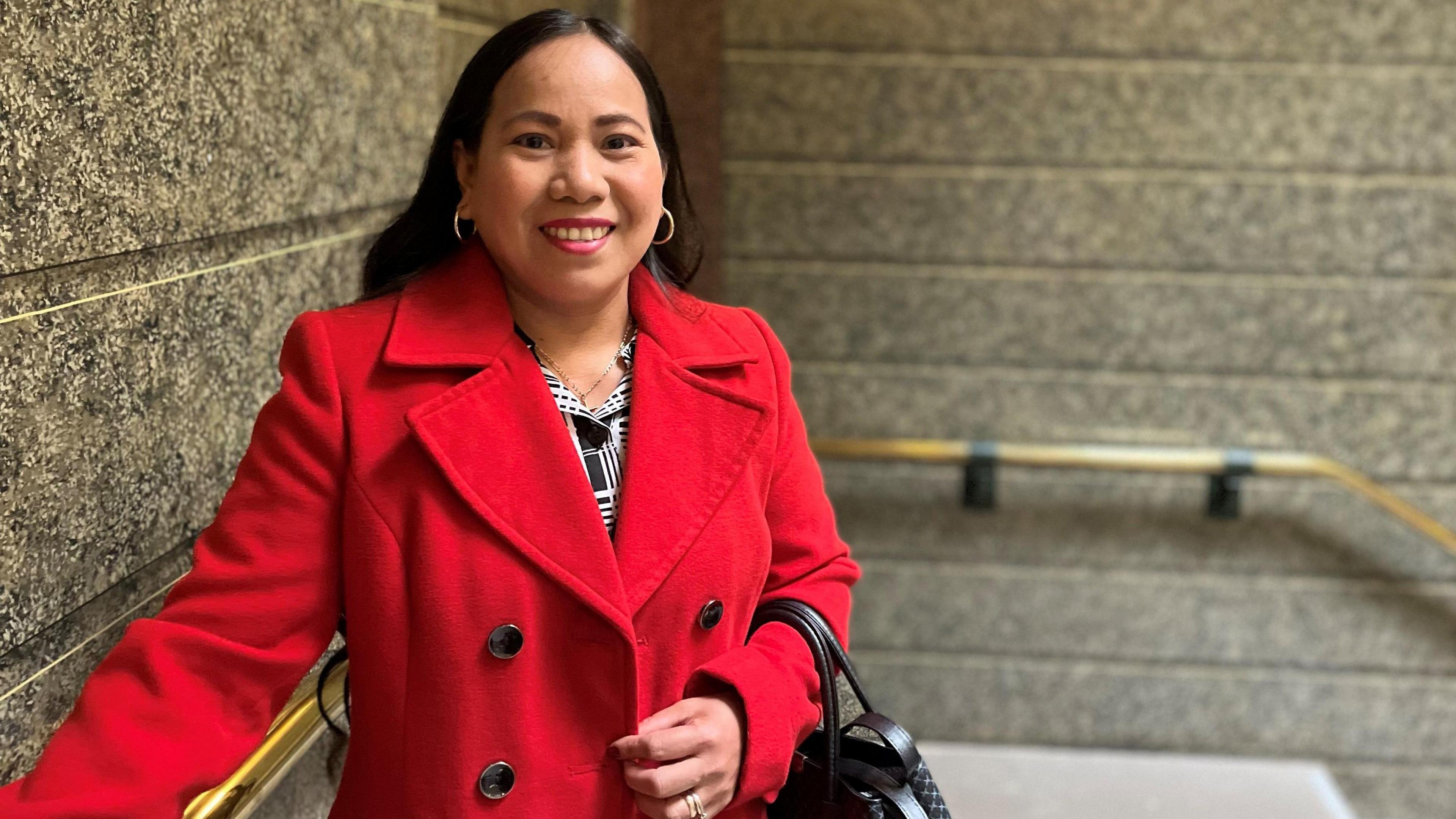 Alma Partington is smiling as she stands next to a sweeping staircase. She has long black hair and is wearing a red coat and a black and white top, with a black handbag over her arm.