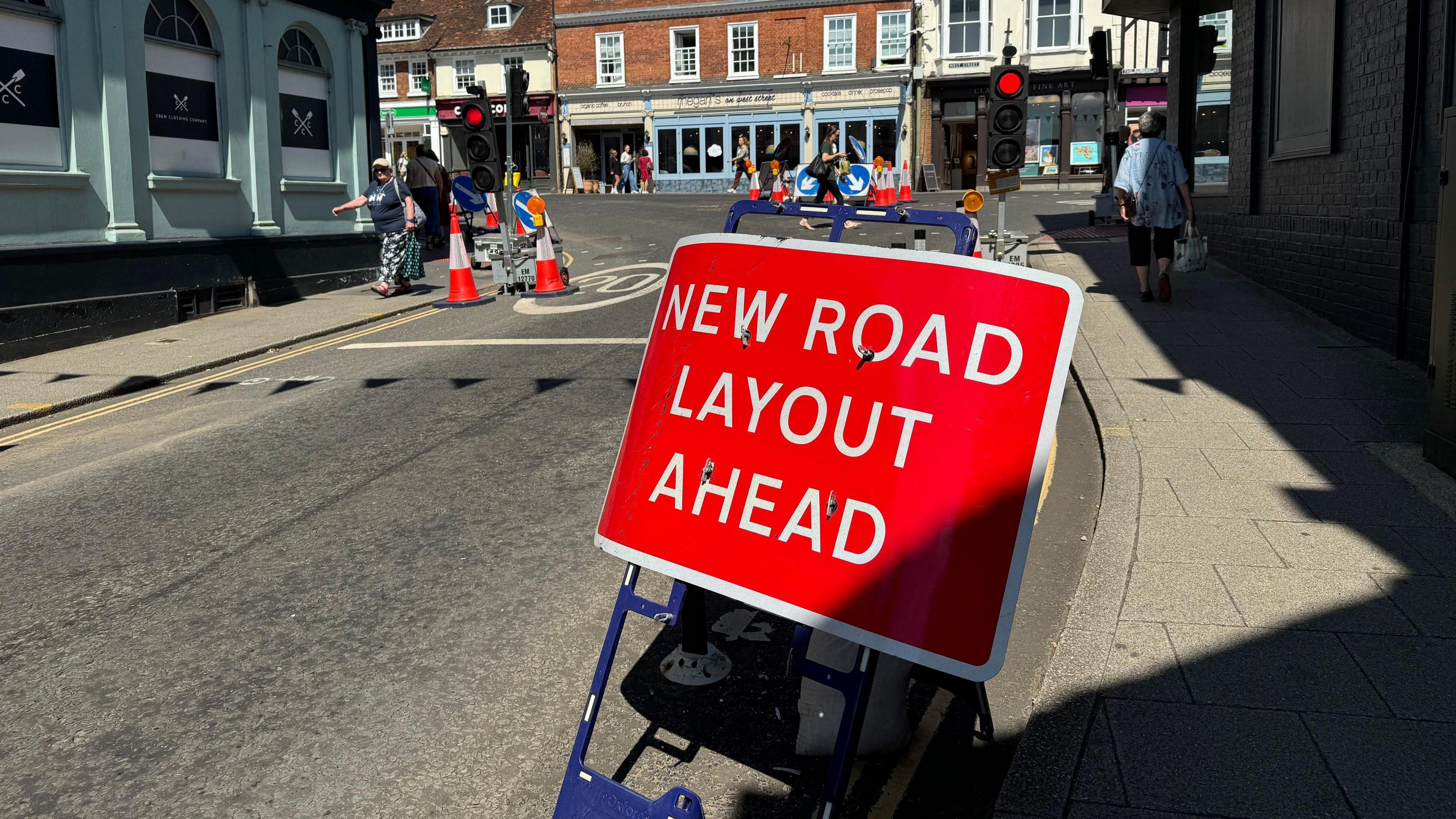 Downing Street in Farnham town centre. A red sign with white letters says: "NEW ROAD LAYOUT AHEAD." Behind it are temporary traffic lights. The light on them is red. The are orange and white traffic cones. Shoppers can be seen in the background, as well as shops on a section of the high street.