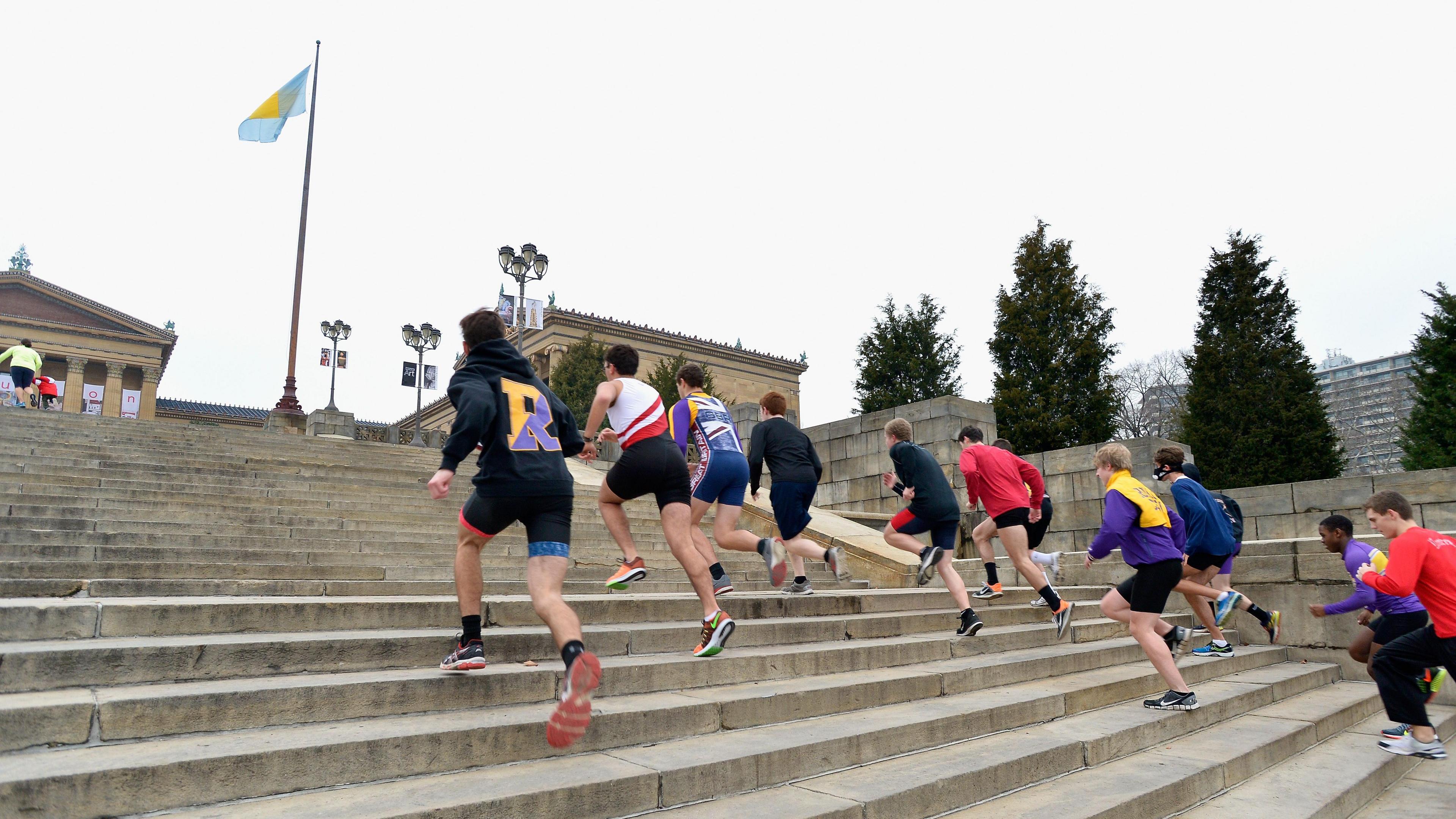 Group of people running up the grey steps of the Philadelphia Museum of Art, many of them are in running kit.  A flag flies from a pole to the left, with the pillars of a stone building visible on the left.