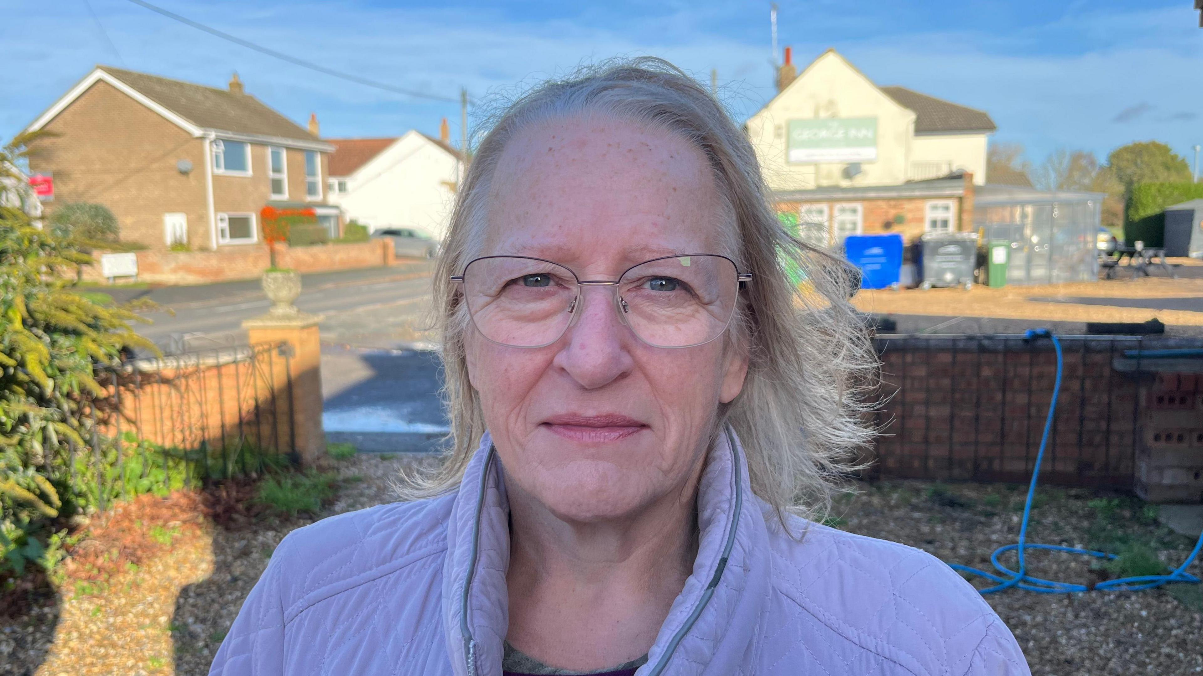 Teresa Coplestone, 63, has long fair hair and glasses and is wearing a lilac padded jacket with a dark top underneath. She is standing in a front garden with a road behind her and a gravel driveway is visible. There are some buildings too, with a house and a pub on the right.
