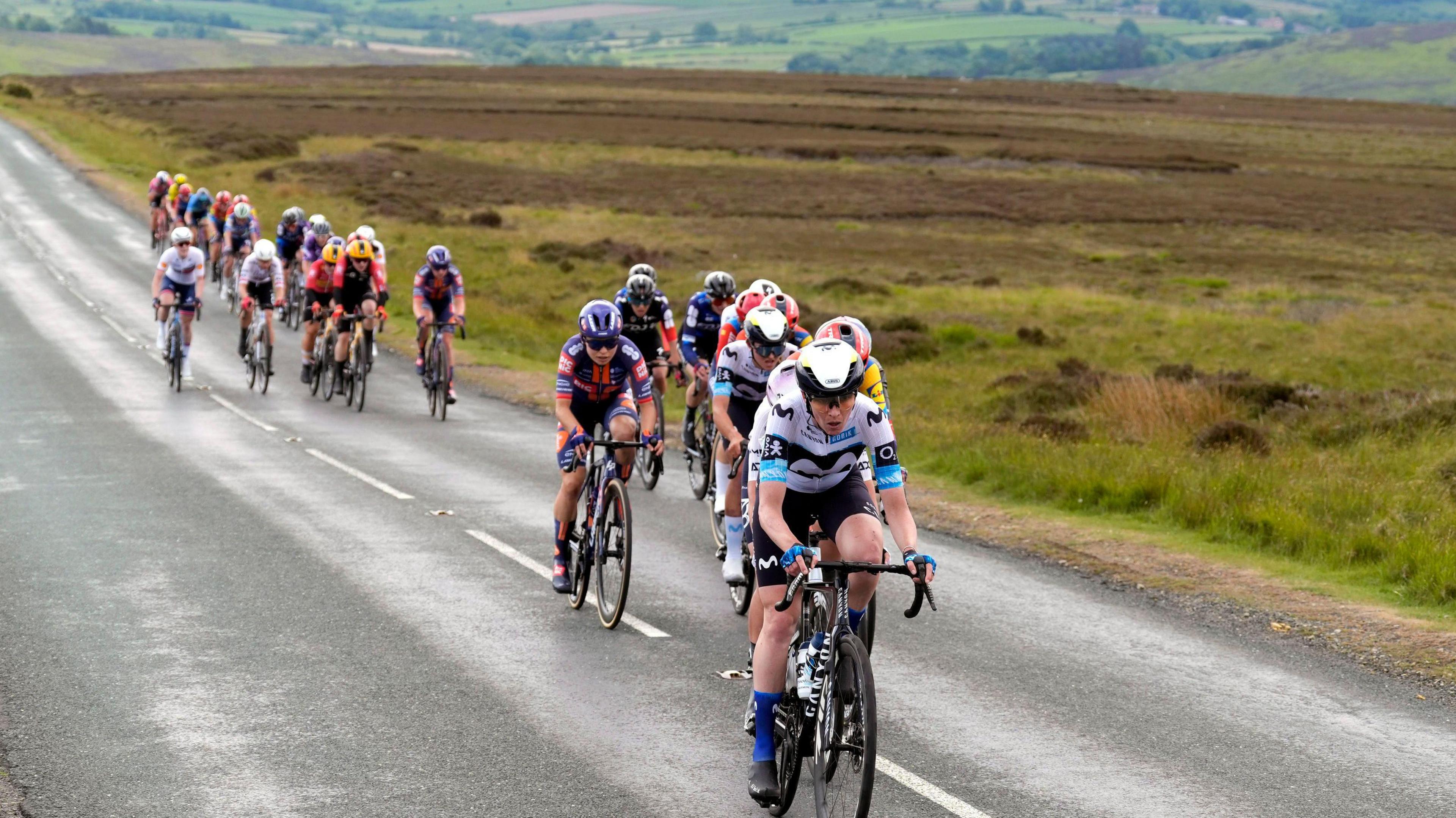 A group of cyclists, all wearing lycra and helmets, riding along a country road. In the background is sprawling heathland.