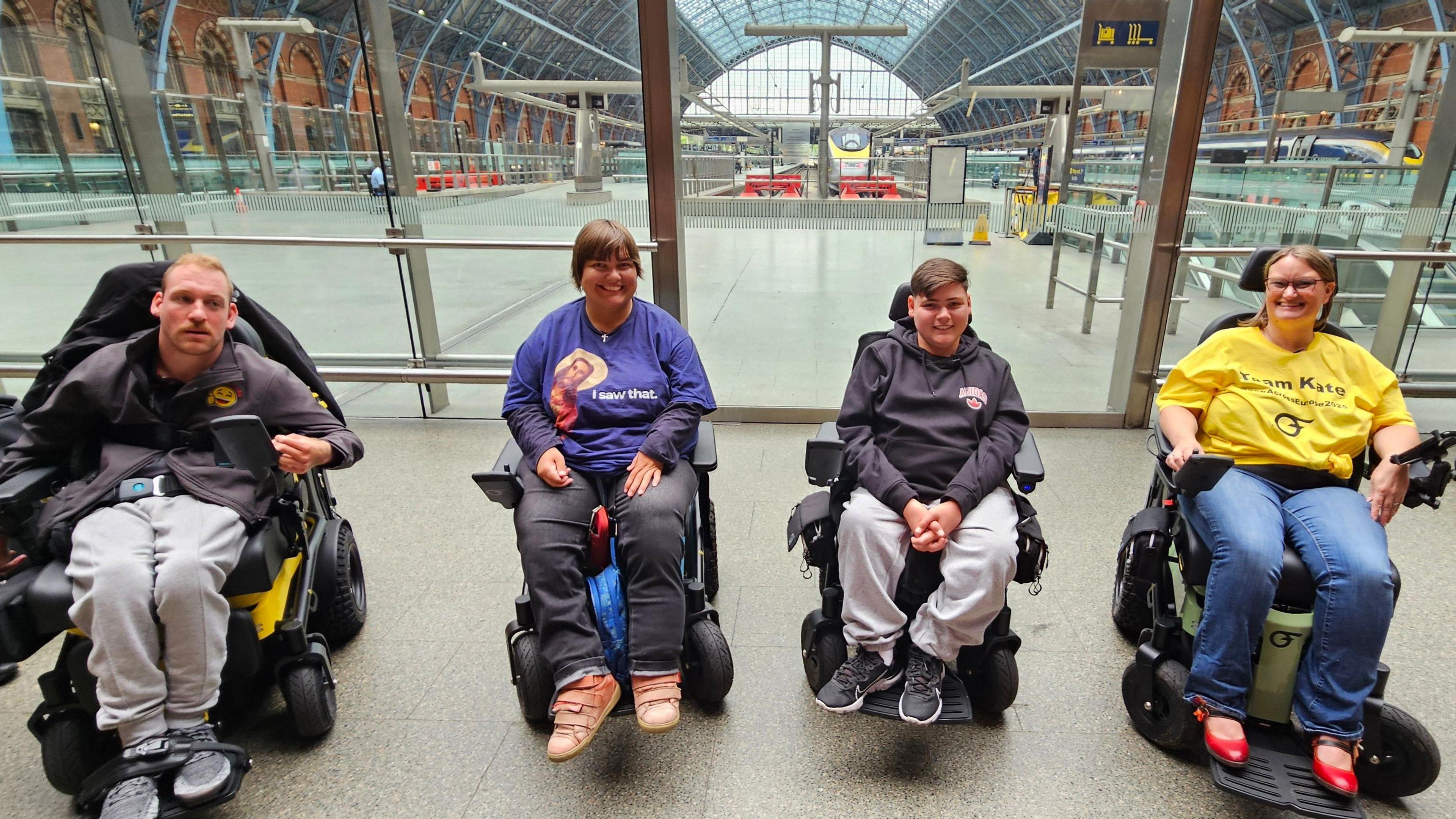 Four people using wheelchairs sat inside a large train station looking at the camera.
