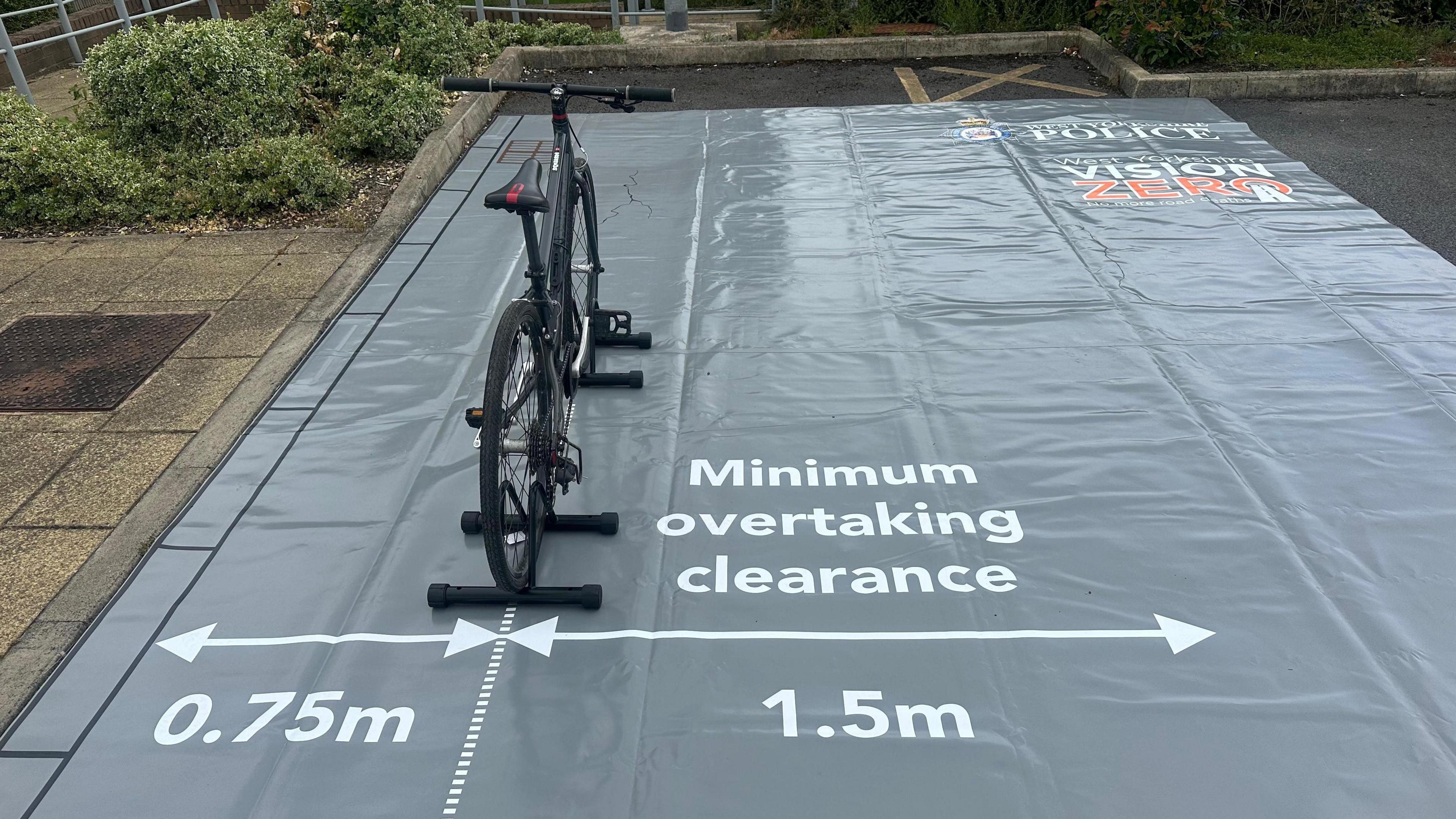 A bicycle on a stand on top of a plastic mat that has arrows indicating 0.75 metres and 1.5 metres which is labelled minimum overtaking clearance.