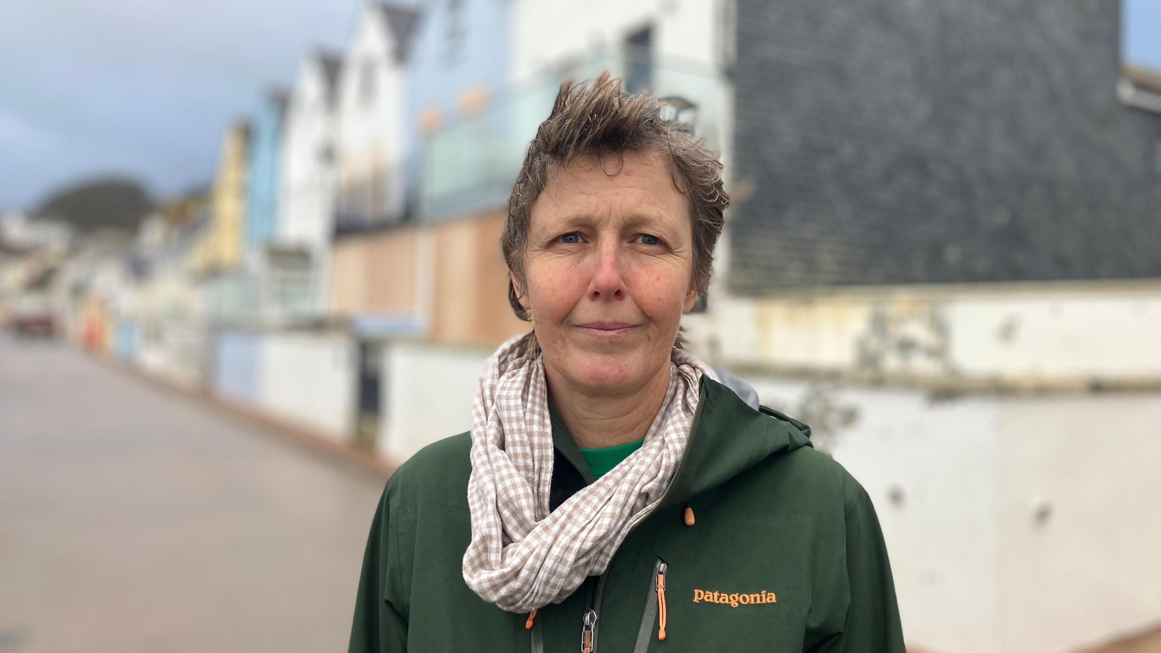 Gail Stubbs stands on the seafront in the seaside town of Torcross in Devon, there is a line of colourful seafront homes in the background, some have windows and doors boarded up. Gail is wearing a green waterproof coat