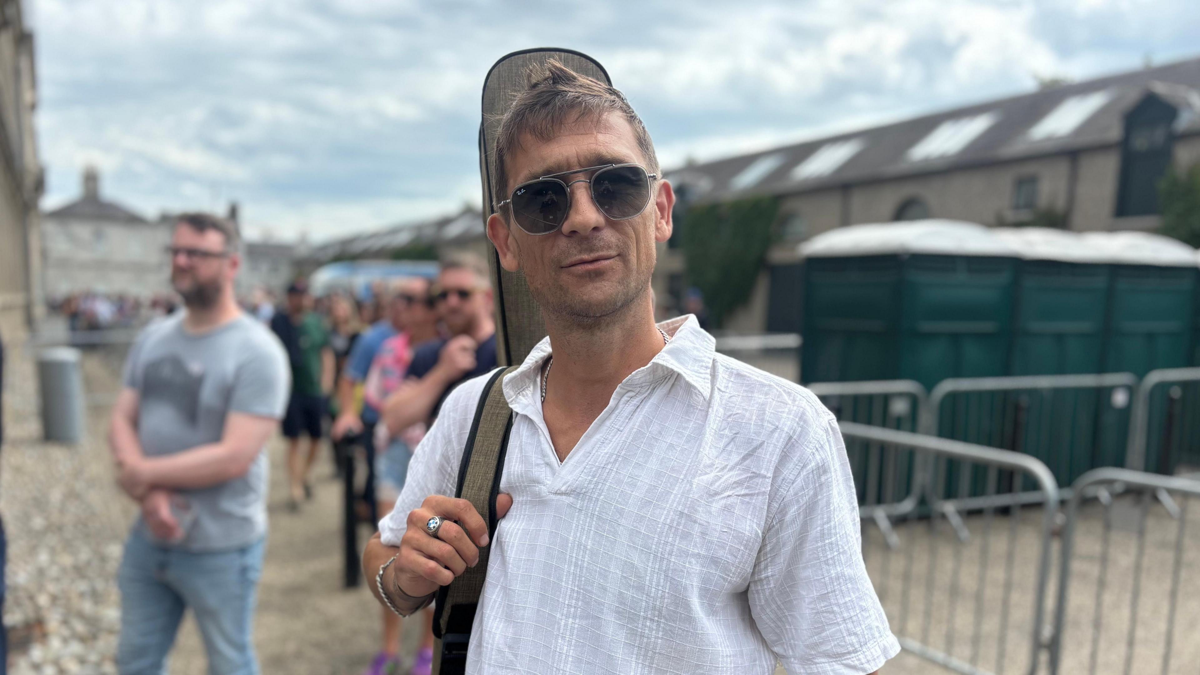 Man with a white t-shirt on and a guitar stands in a queue, he has dark hair and sunglasses