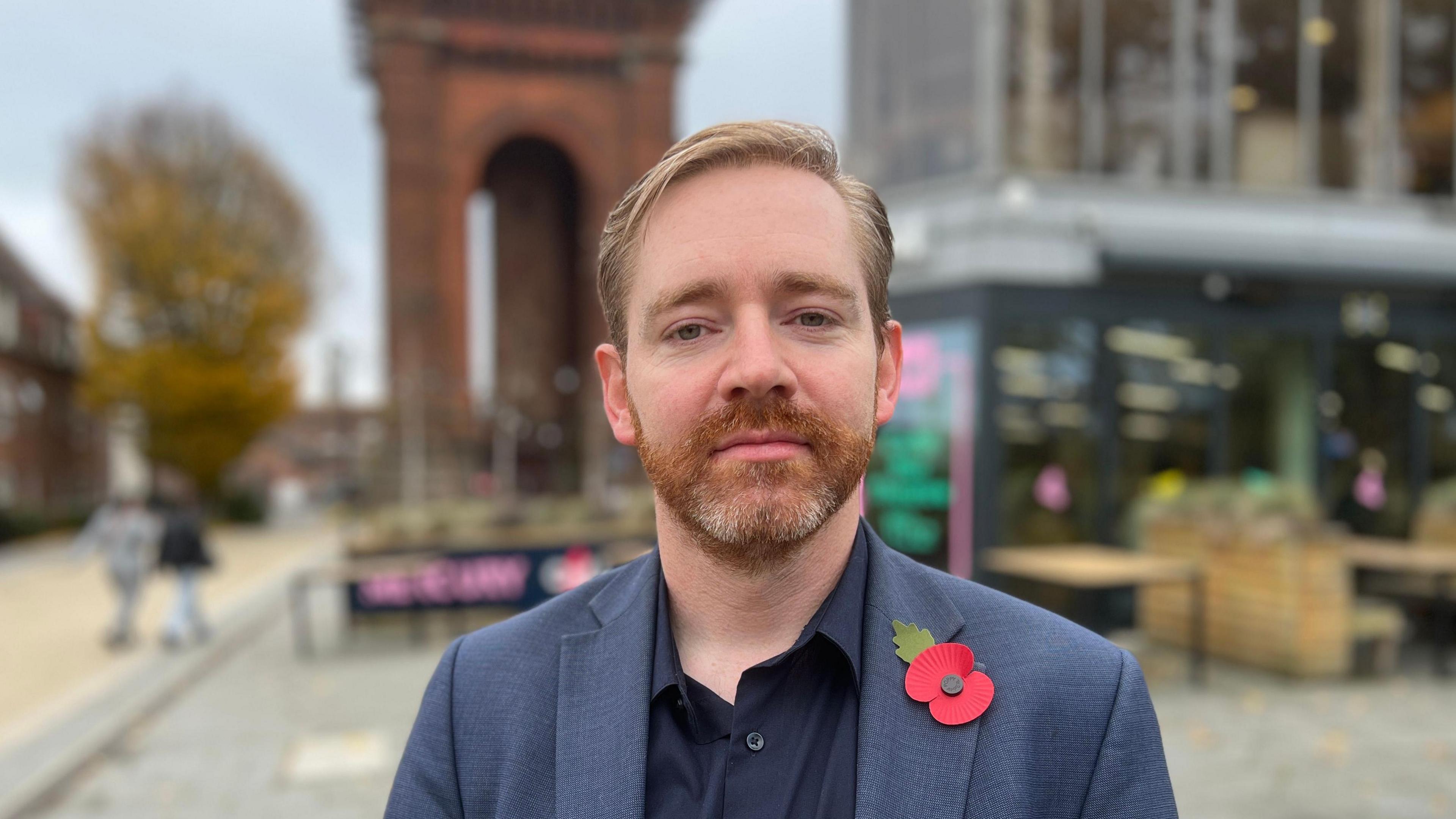 Adam Fox stands in Colchester city centre which is blurred behind him. He slightly smiles at the camera. He has light hair and a red and grey beard. He wears a dark blue suit jacket with a red poppy on one side of his chest and a black shirt underneath.