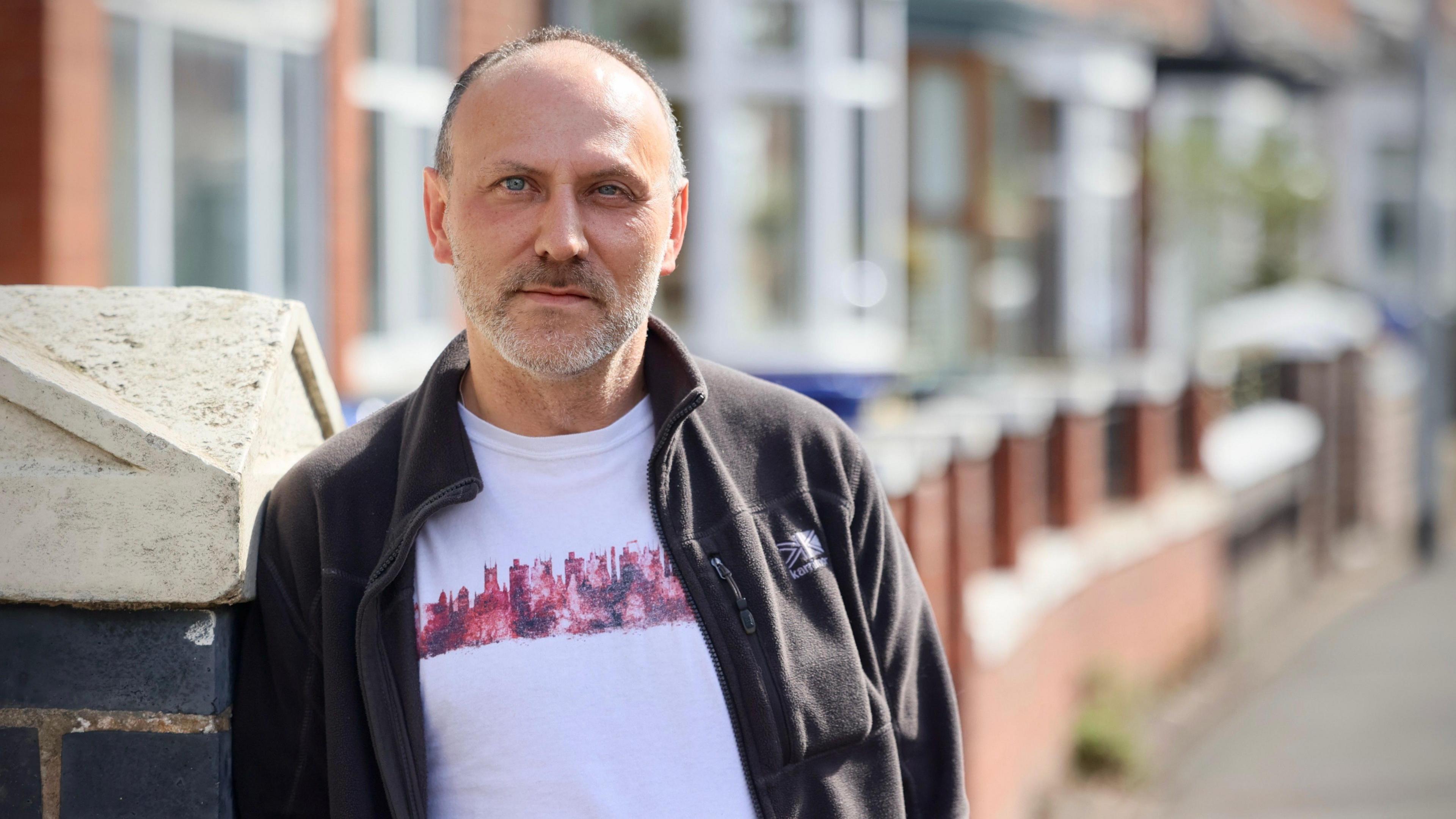 Andy Polshaw, a man wearing a white t-shirt and a black fleece jacket is standing on a street with terraced houses behind him.