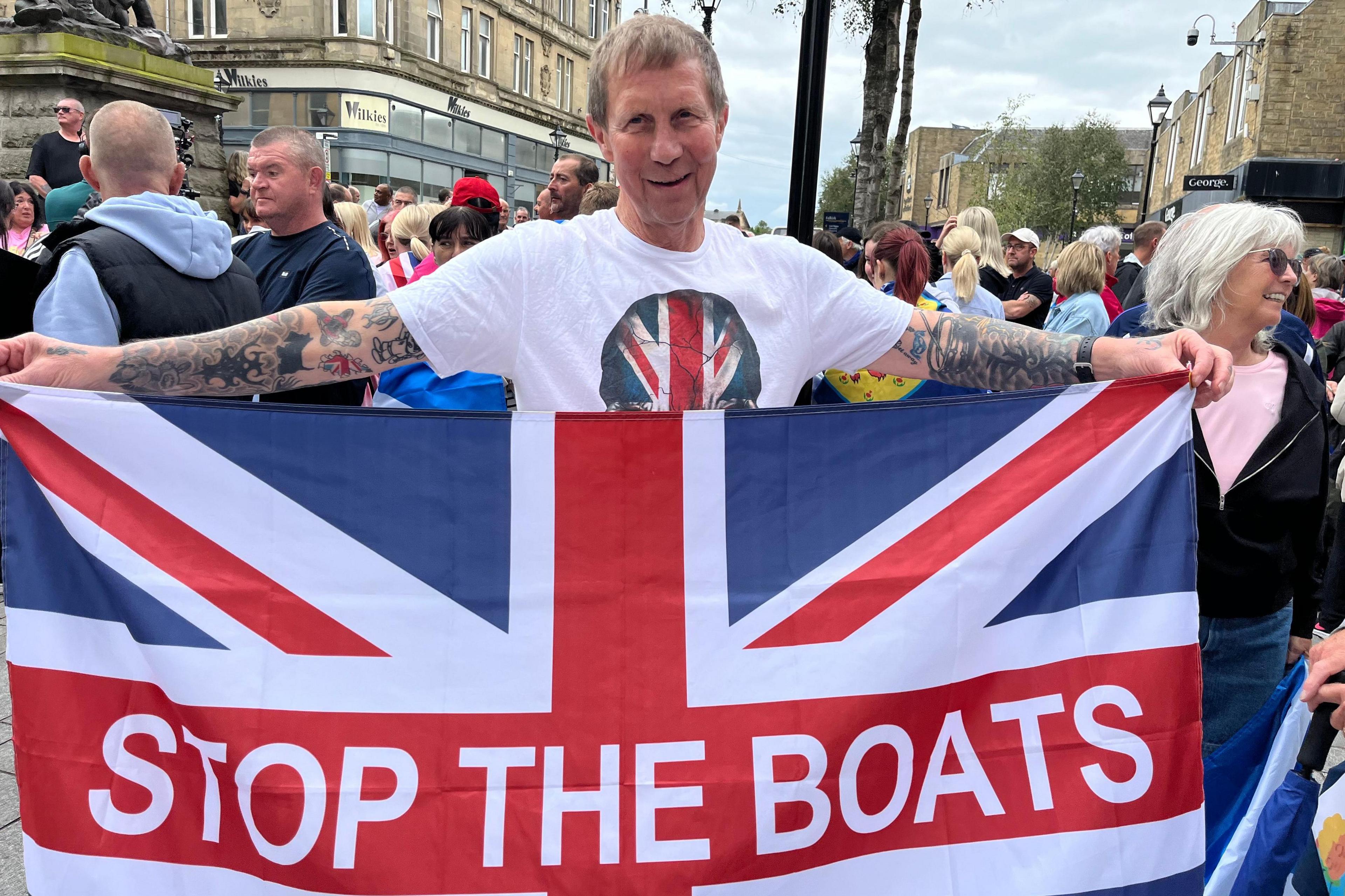 A man in a white T-shirt with short brown hair holds a union flag with Stop the Boats written on it in white letters.