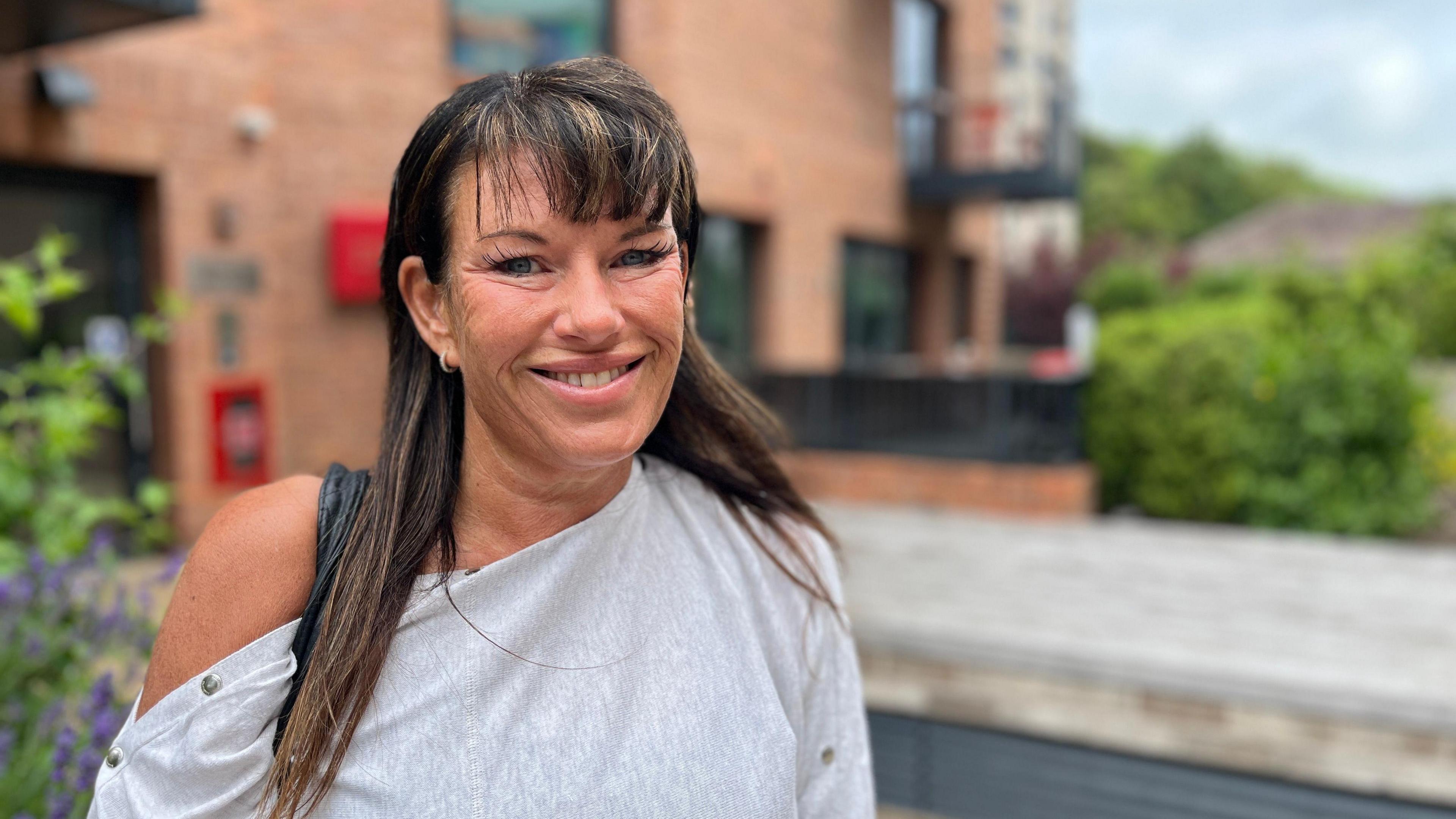 A picture of social housing tenant, Mary Davies, in Brighton. She looks directly at the camera and is smiling. She is wearing a grey top and has long dark hair with a fringe. An apartment block can be seen in the background.