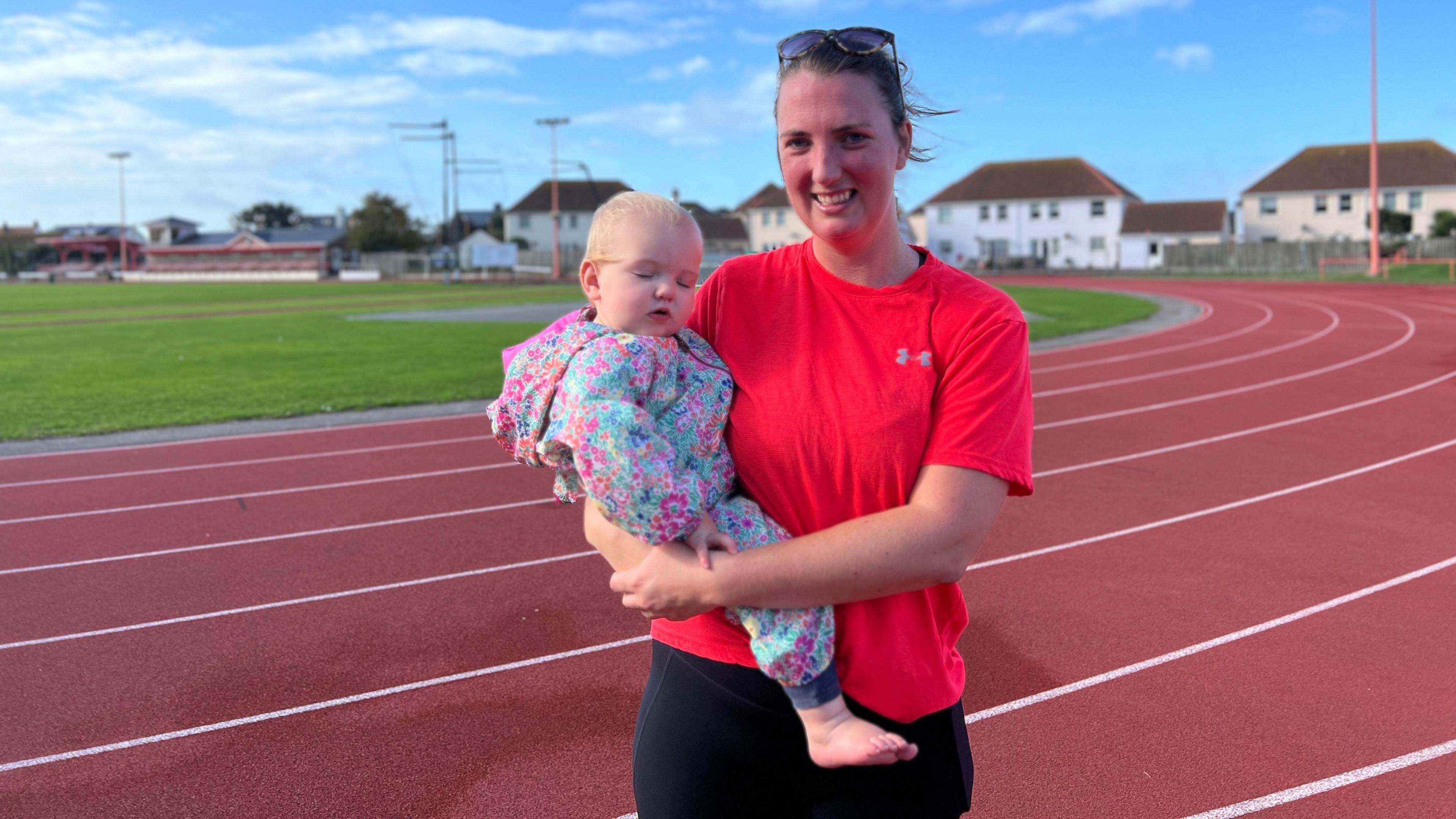 Lisa smiles at the camera with the running track behind her and the sky is blue. She's wearing a red exercise top with black joggers and she has glasses on the top of her head. Her baby is a bright all-in-one with floral patterns and has her eyes closed. Lisa is a bit red in the face after the running session.