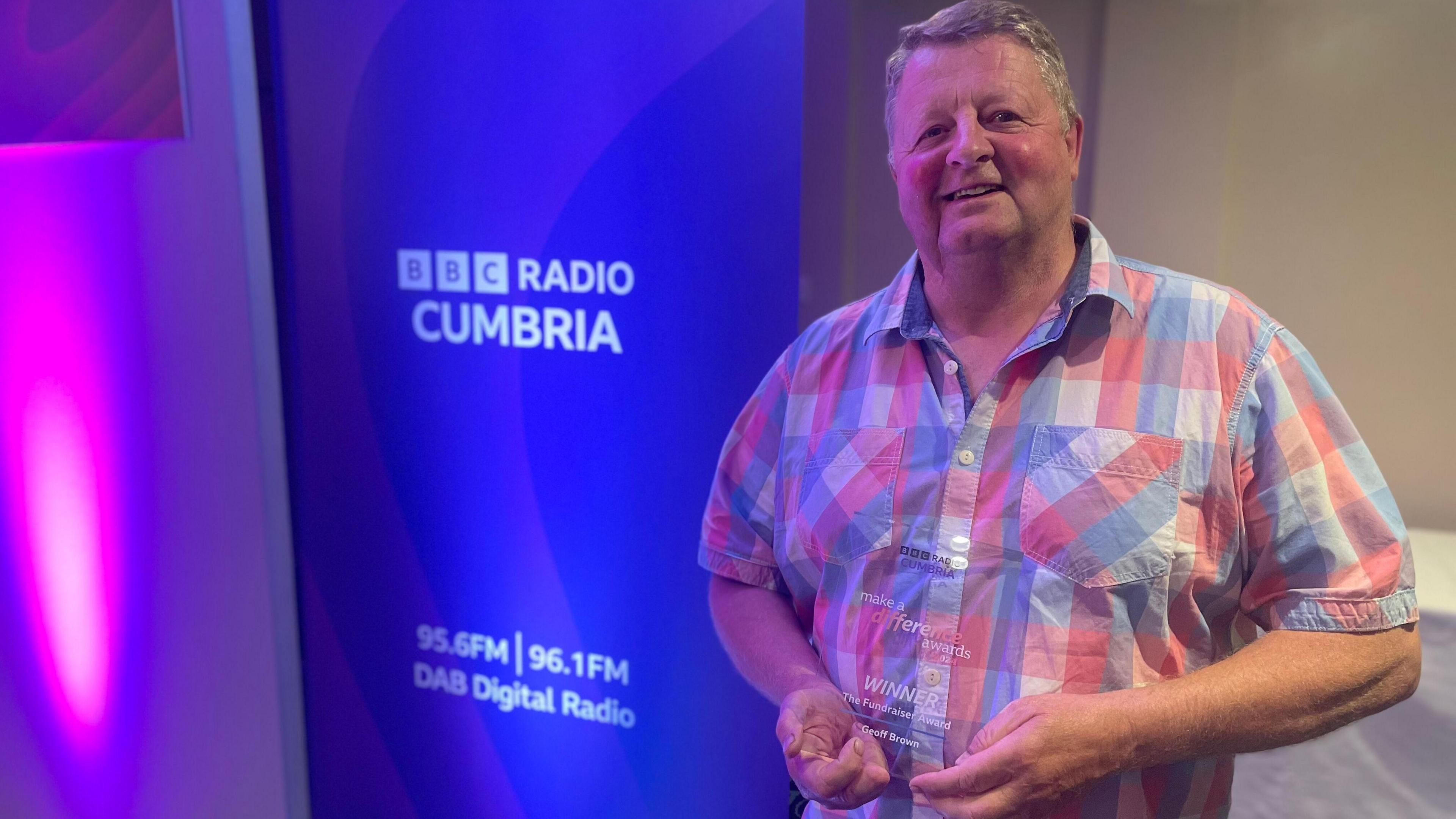 Geoff standing with his fundraiser award in front of purple BBC Radio Cumbria signage.