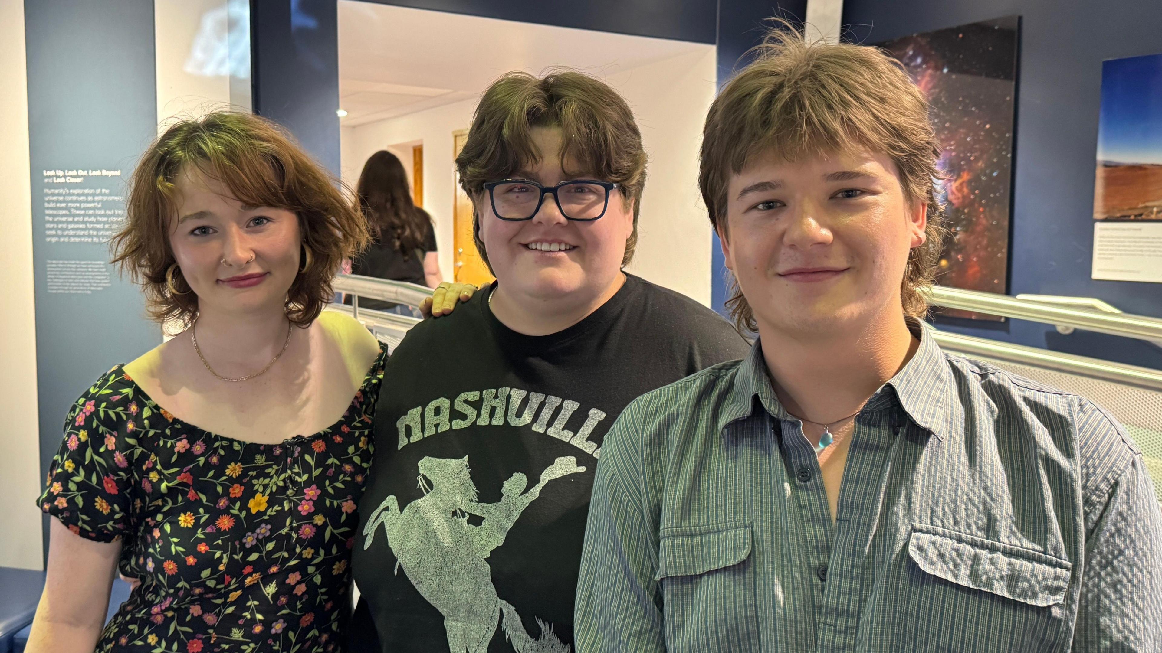 Three friends - Emma, Bec and Arron - stand side by side smiling at the camera. Emma on the left has short hair and is wearing a floral dress. Bec in the middle has short, dark hair and is wearing a pair of square glasses and a black t-shirt. Arron on the right has longer hair and is wearing a stripe shirt and a necklace.