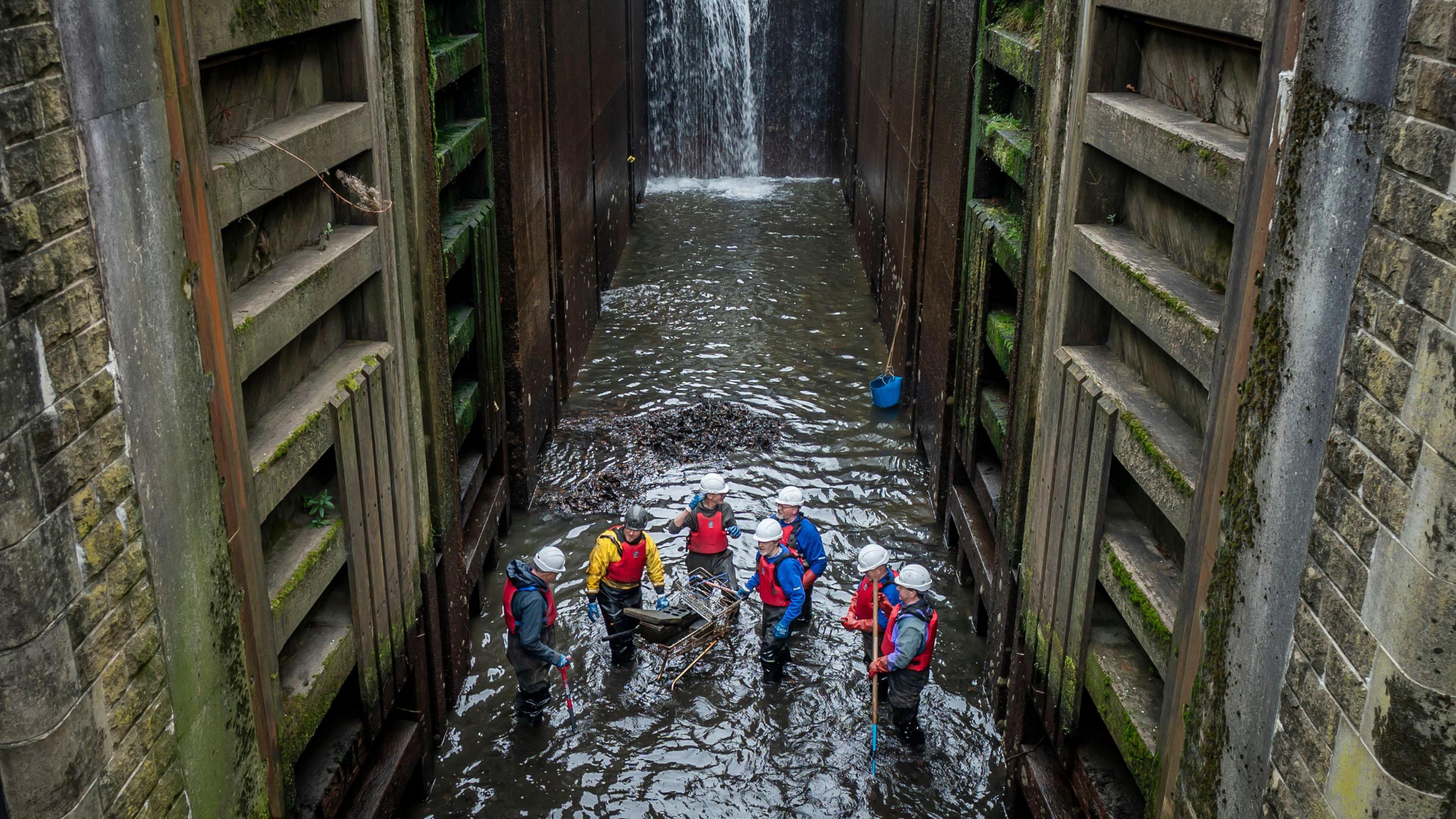 A group of seven volunteers stand around a shopping trolley which is partially submerged in the water. The trolley contains large waste items.