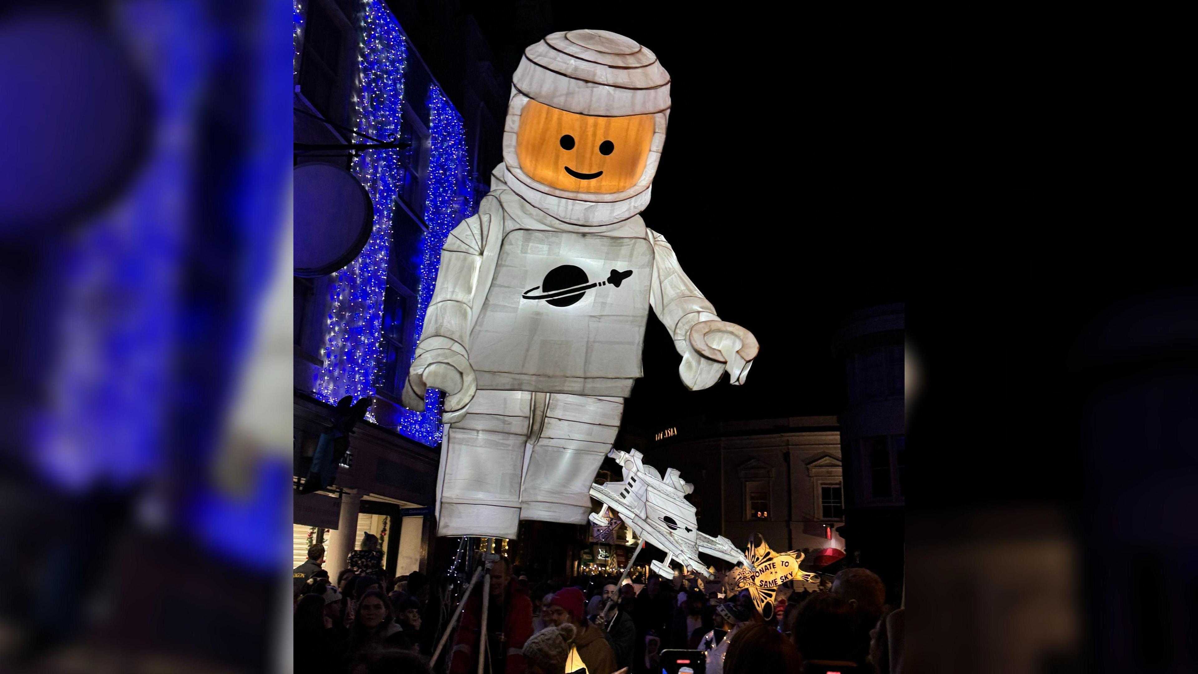 The large Lego astronaut is seen carried above a crowd of people and there are smaller lanterns in the background, in the shape of a spaceship. There are blue fairy lights strung on a building and people lining the streets 