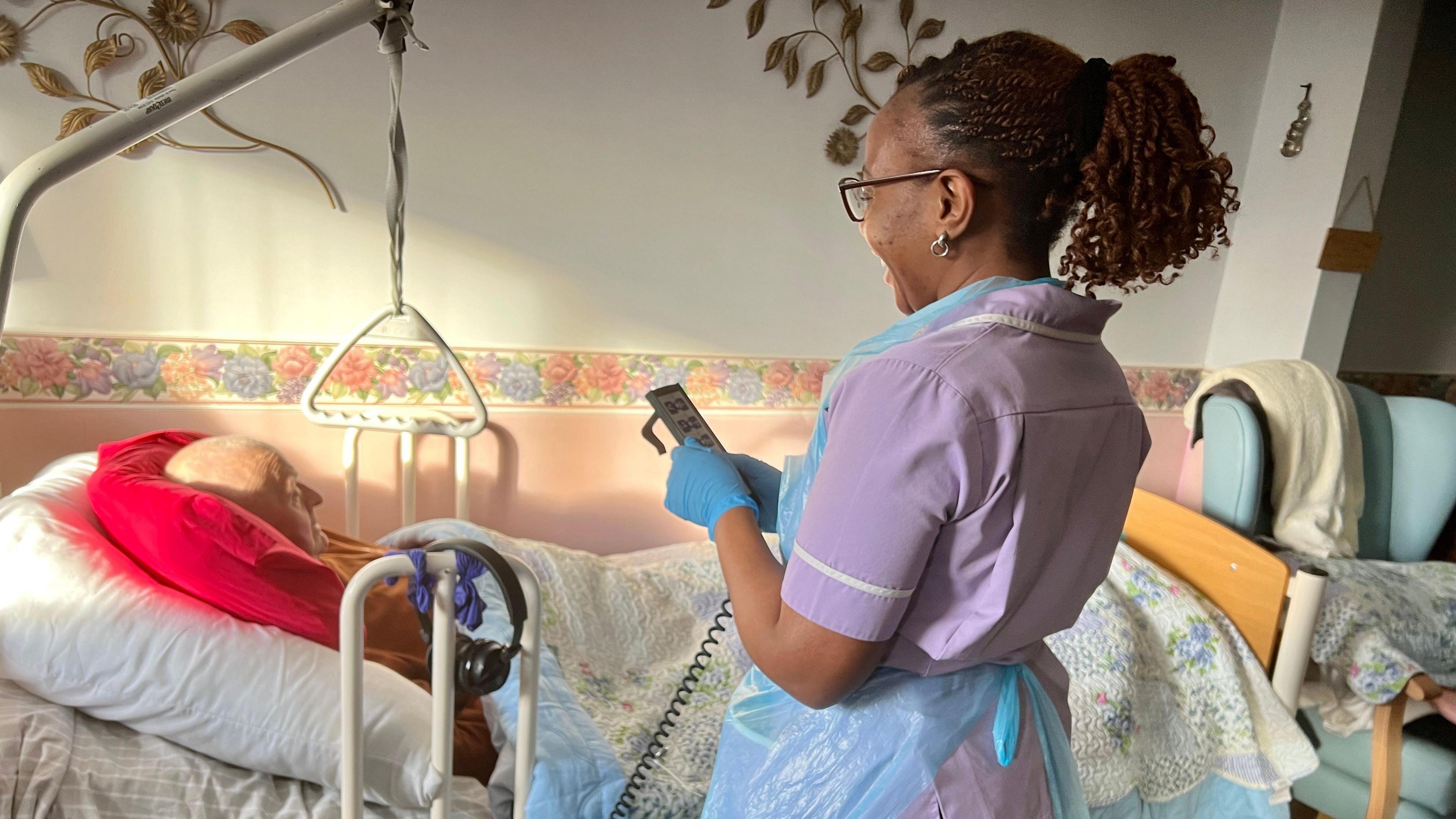 Derek lying in his bed in his living room being cared for by a female carer from Primary Homecare in Suffolk. She is wearing a purple uniform and latex gloves. She is holding the control for the bed, trying to make Derek comfortable