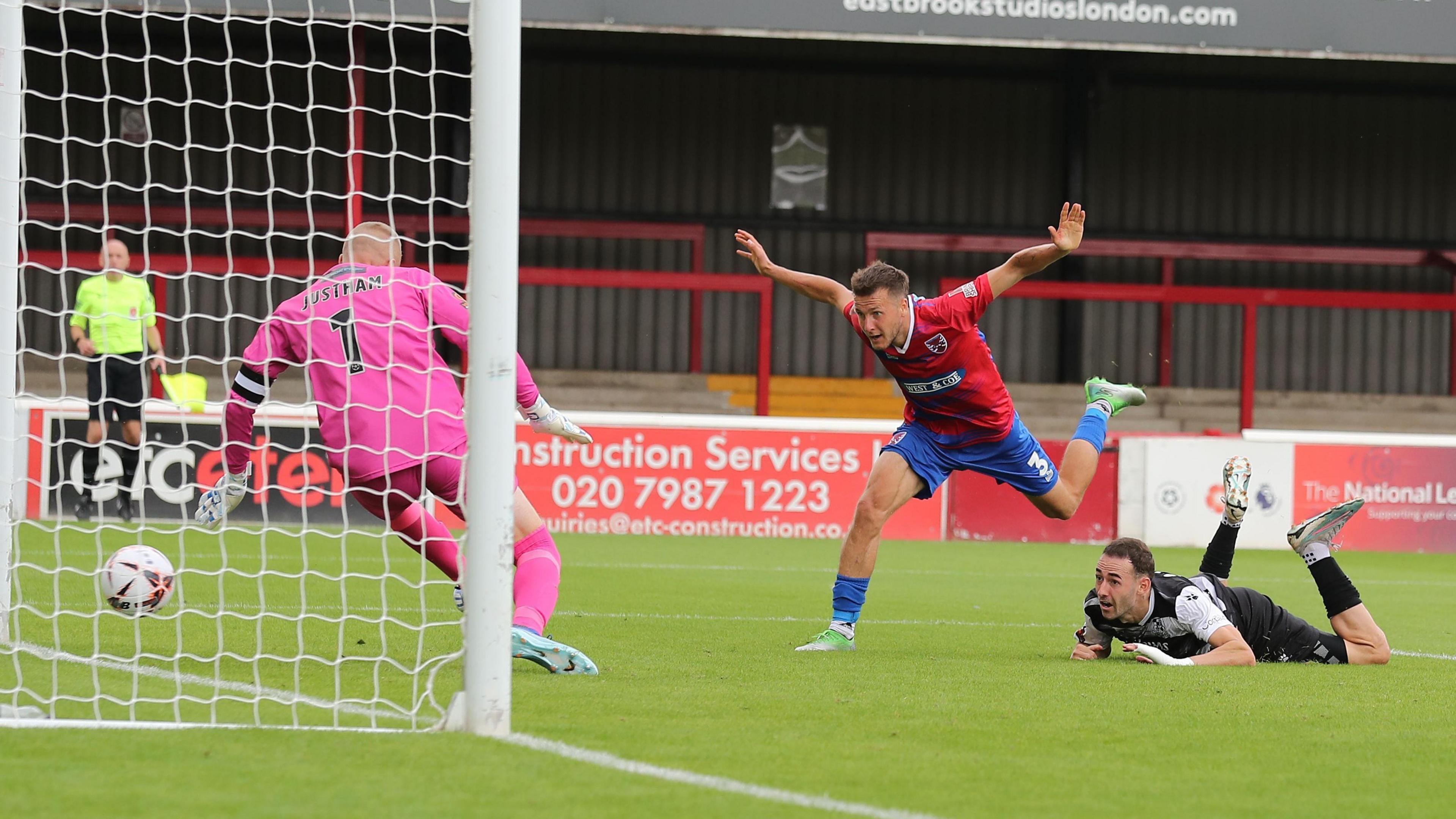 Sam Ashford stumbles off balance as his shot goes beyond the Wealdstone goalkeeper into the net for a Woking goal, with a defender lying on the ball next to him after attempting to make a tackle