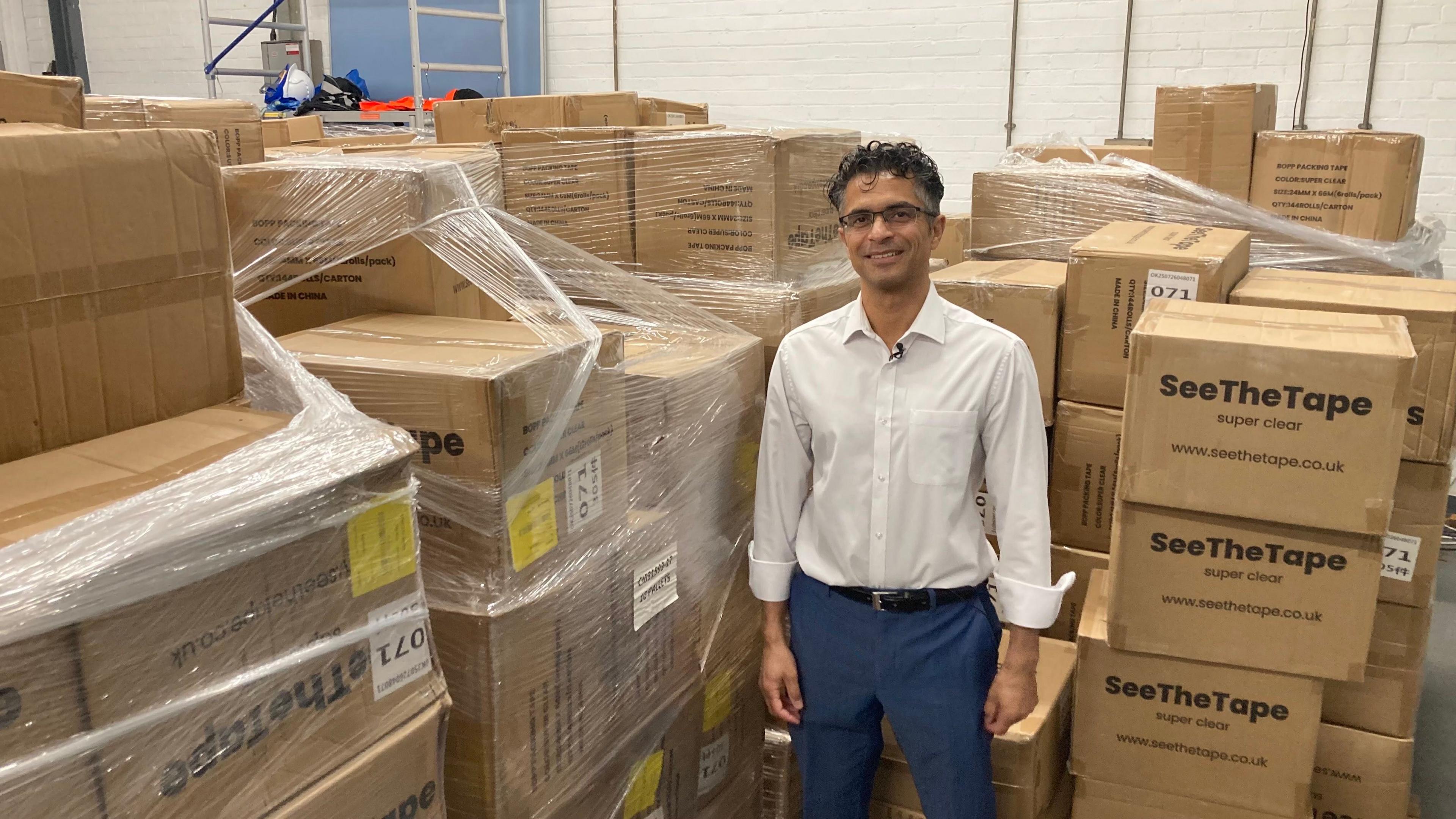Dr Rajan Bhojwani standing in front of stacks of boxes in a warehouse. The boxes display the brand name See The Tape.