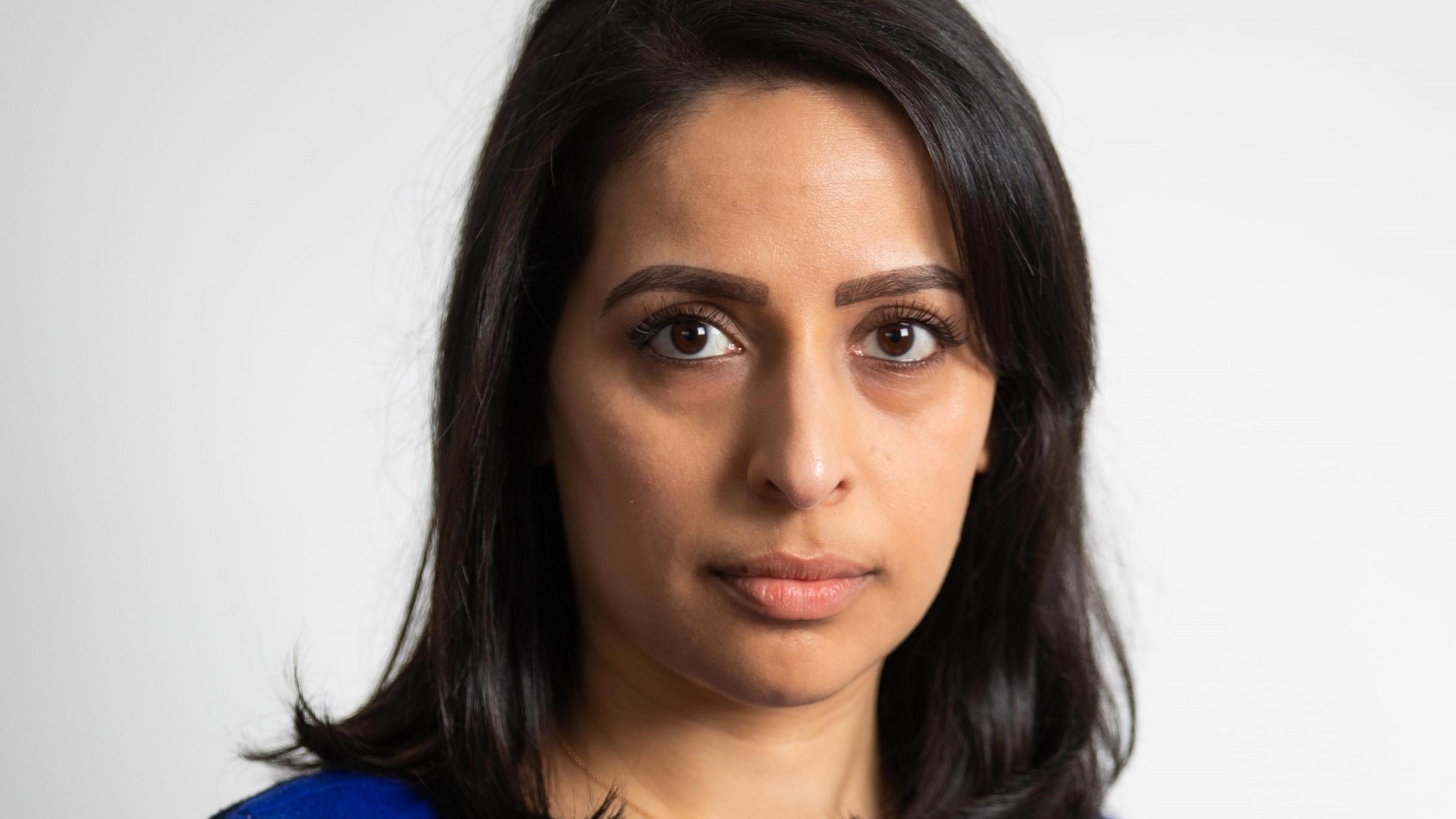 Dr Patel is looking directly at the camera. She has shoulder length dark hair and is wearing a bright blue top. She is in front of a grey background.