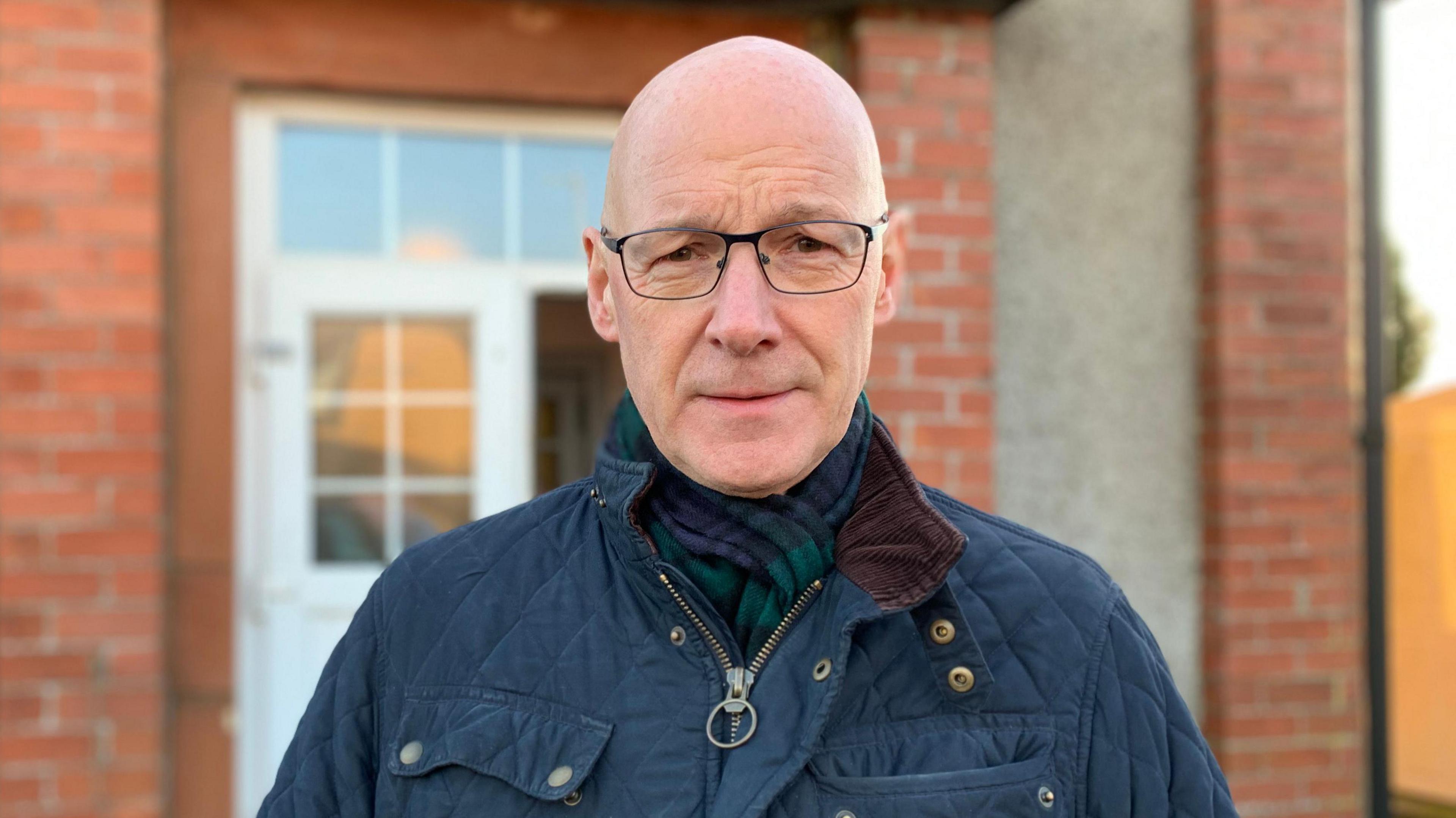 A bald headed man in glasses wearing a dark blue coat and a tartan scarf stands in front of a brick village hall building