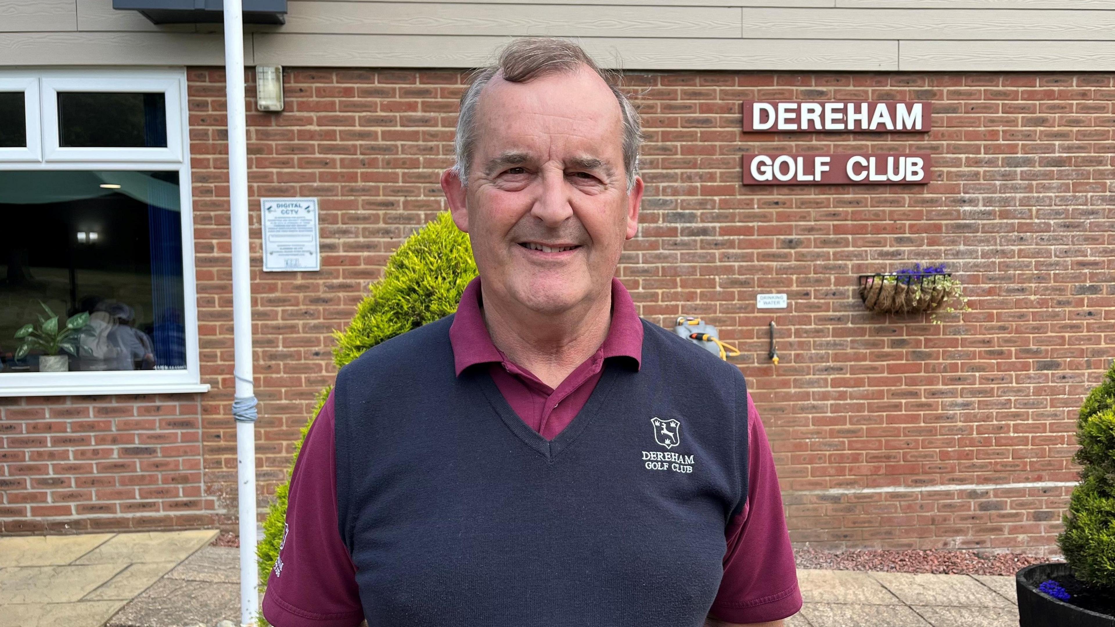 Stuart Goodman, wearing a purple shirt and navy jumper, is standing in front of the clubhouse, with a sign on the wall behind that reads Dereham Golf Club.