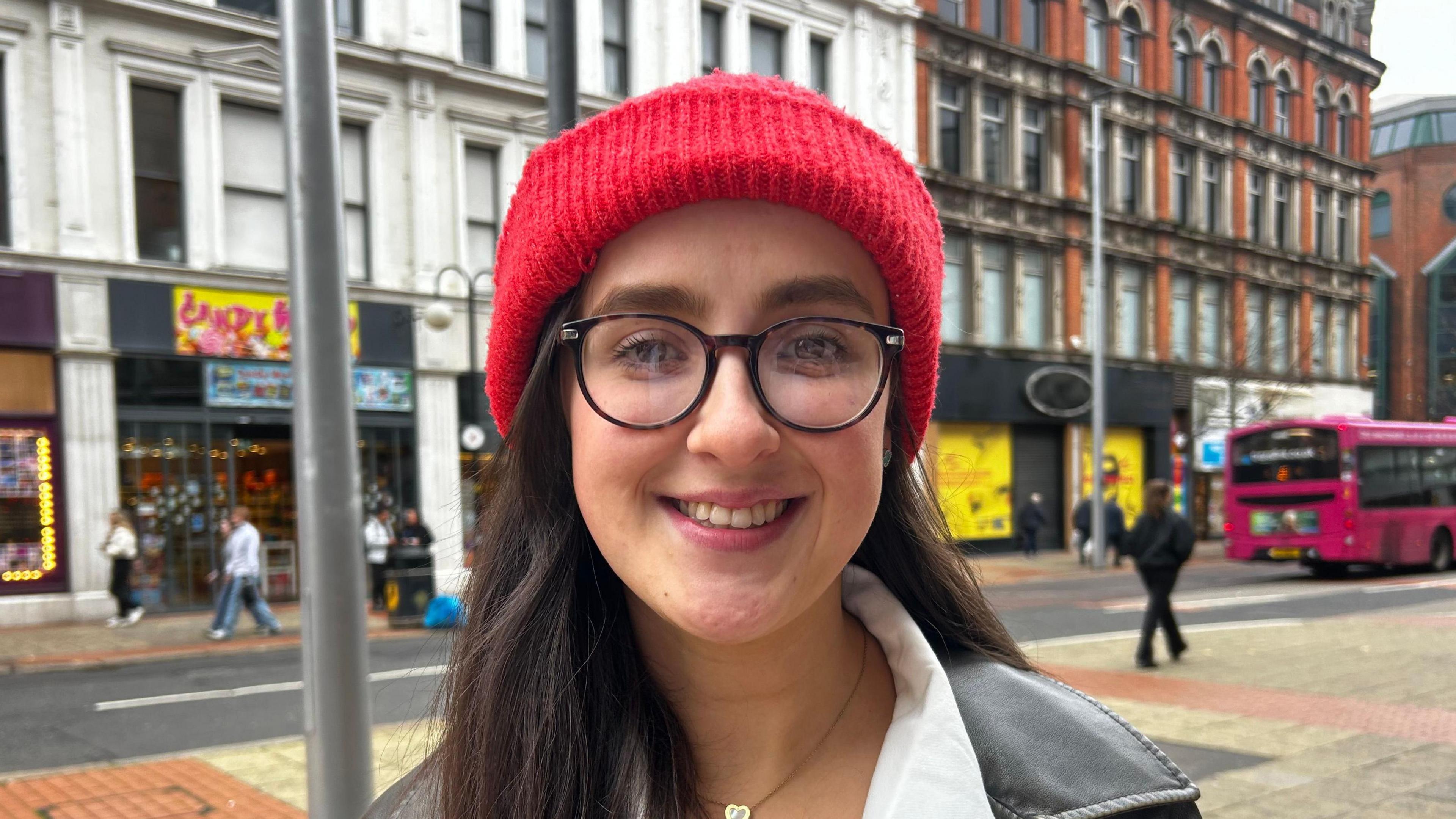 A person with long dark hair and glasses. The are wearing a red beanie hat. They are smiling at the camera. In the background, there is a pink bus. The appear to be standing on a city street.