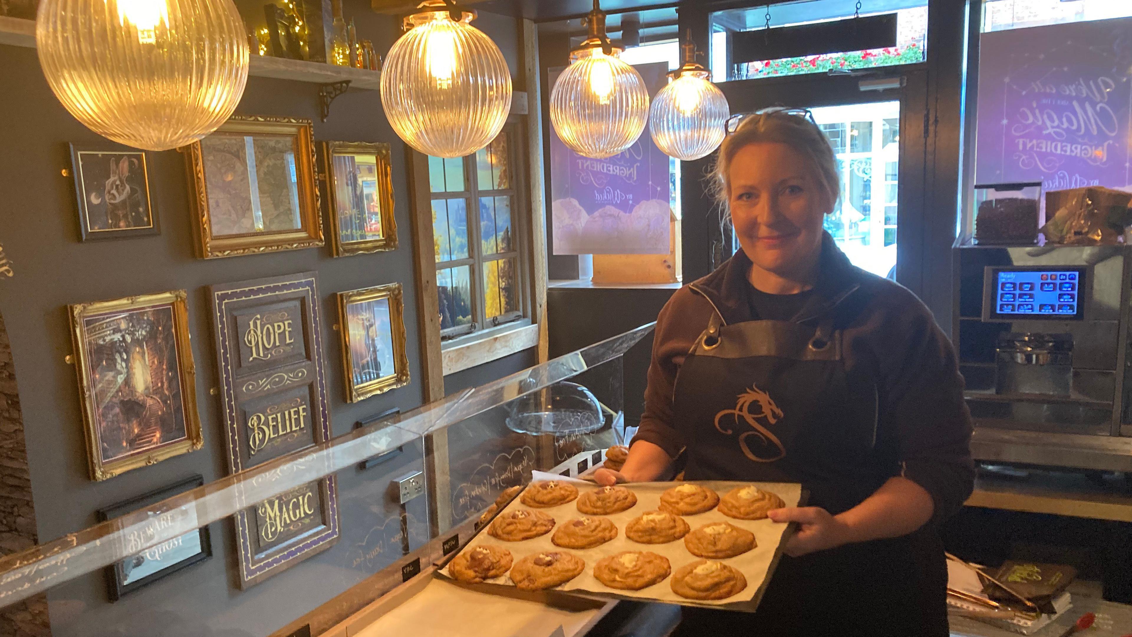 Sally Rees wearing an apron holding a tray of 12 cookies in a shop with magical themed pictures hung on the walls