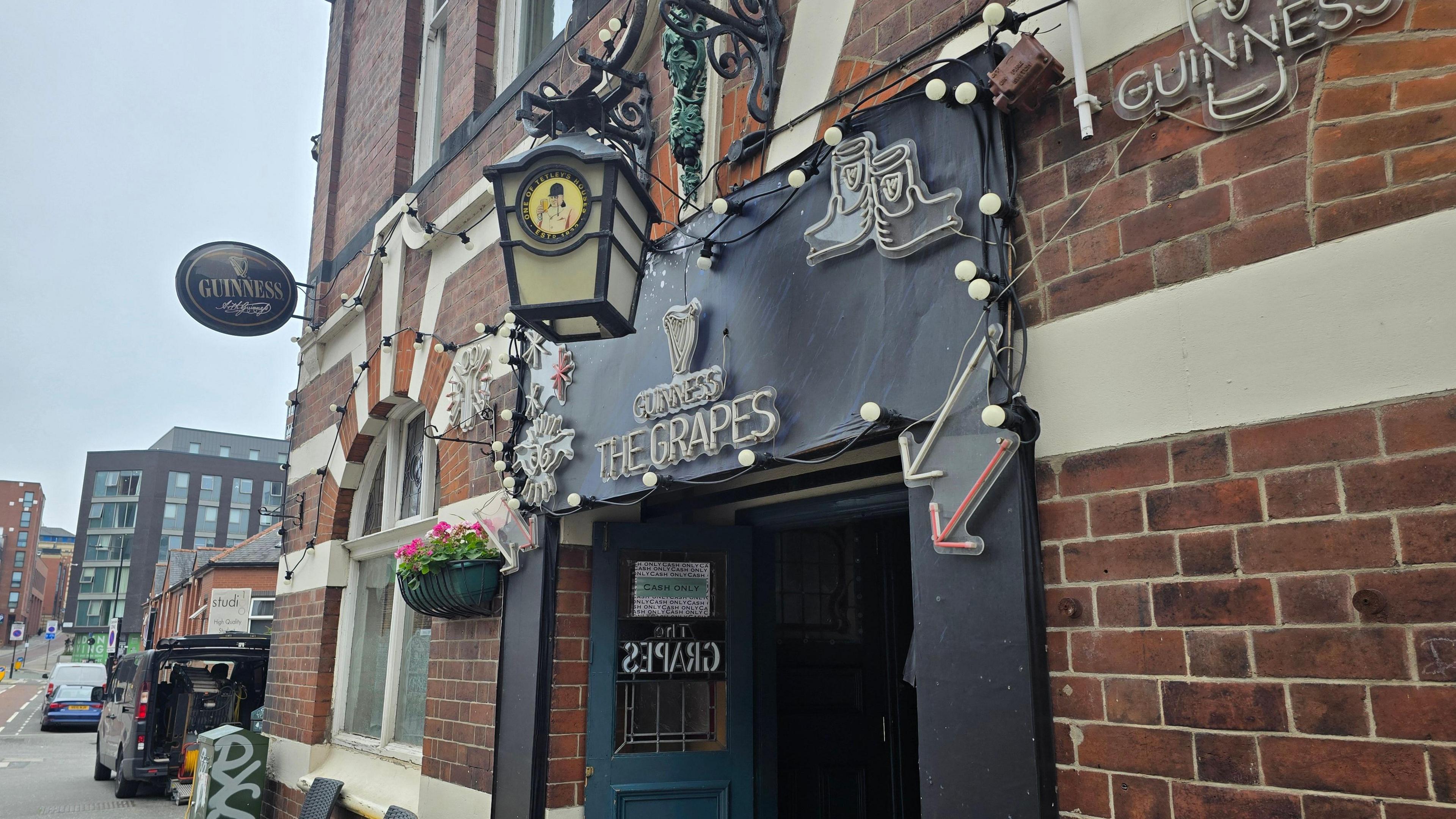 Exterior of 'The Grapes' pub, a red-brick building with white trim. A black sign with white lettering and a pint glass graphic reads 'Guinness' and 'The Grapes.' The entrance features a blue door, hanging flower pots, string lights, and multiple Guinness signs, creating a welcoming pub atmosphere.
