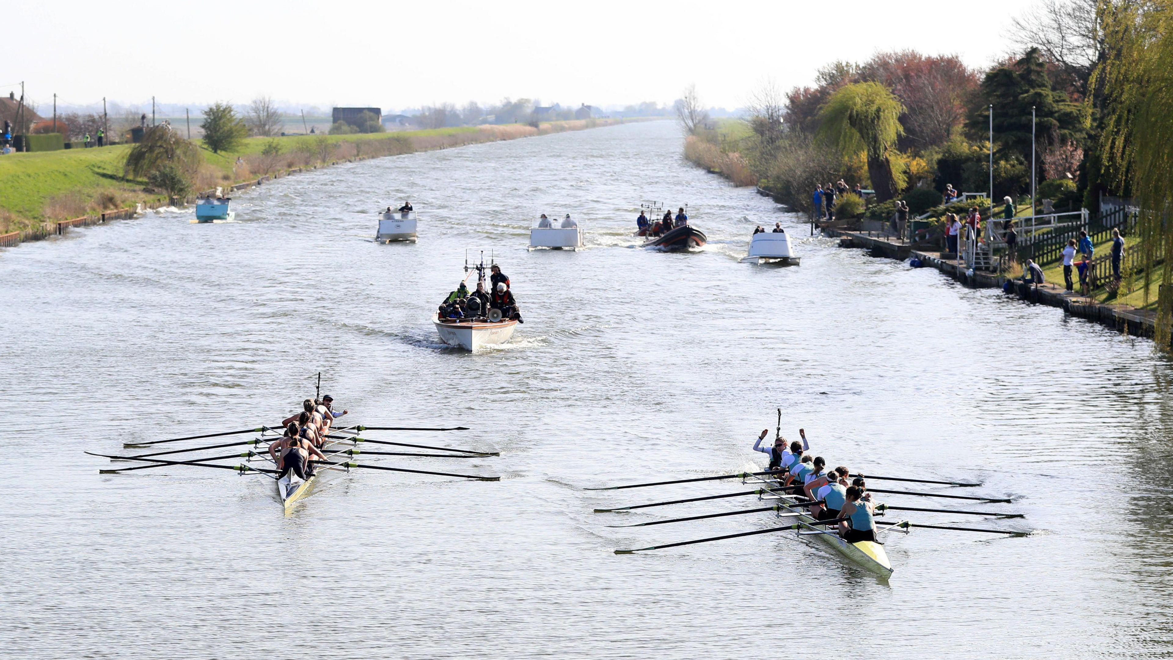 A wide river with two rowing boats of eight people and a cox on either side. Six other support boats are behind them. Spectators stand at the end of gardens on the right-hand side of the riverbank. 