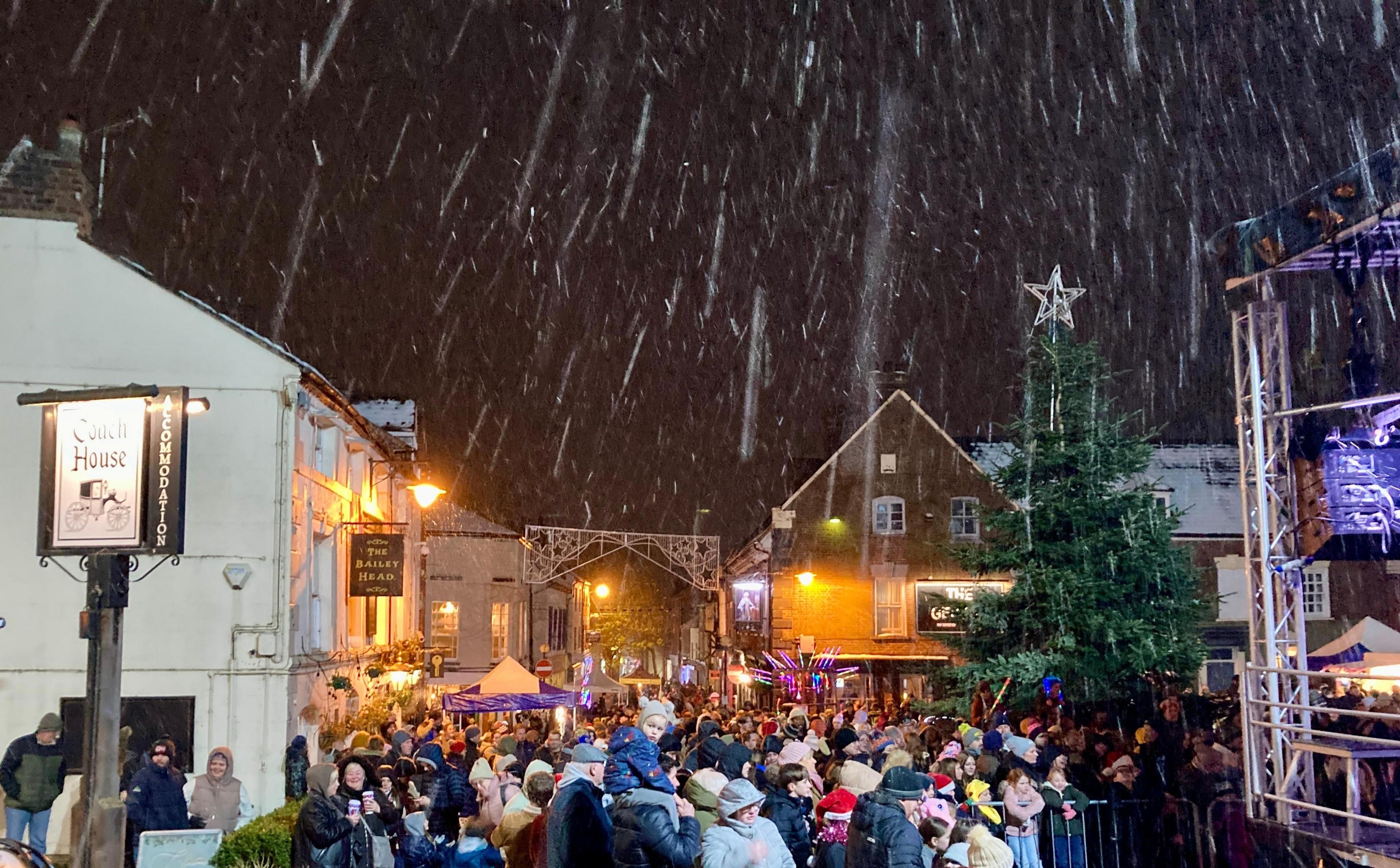 Snow falls on a town square with a crowd of people illuminated by street lights and there are two pub signs on the left - The Coach House and The Bailey Head.  To the right, a metal lattice lighting gantry is visible at the edge of a stage. Christmas lights on a tree and over the street have still to be switched on. 