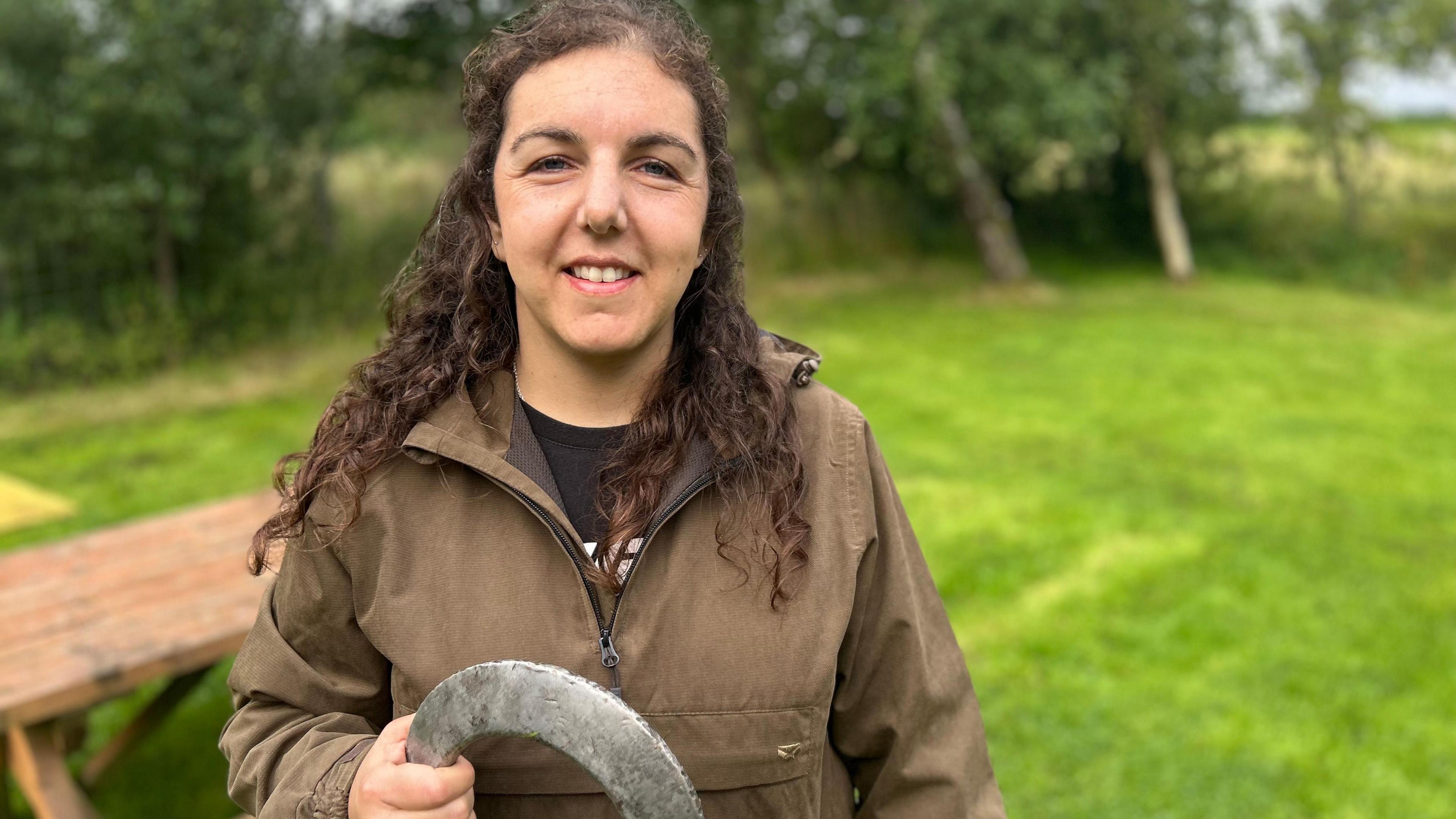 Helen Harkness is smiling at the camera as she holds a quoit. Her curly brown hair is half up half down. She has blue eyes and is wearing a khaki jacket and a black t-shirt.