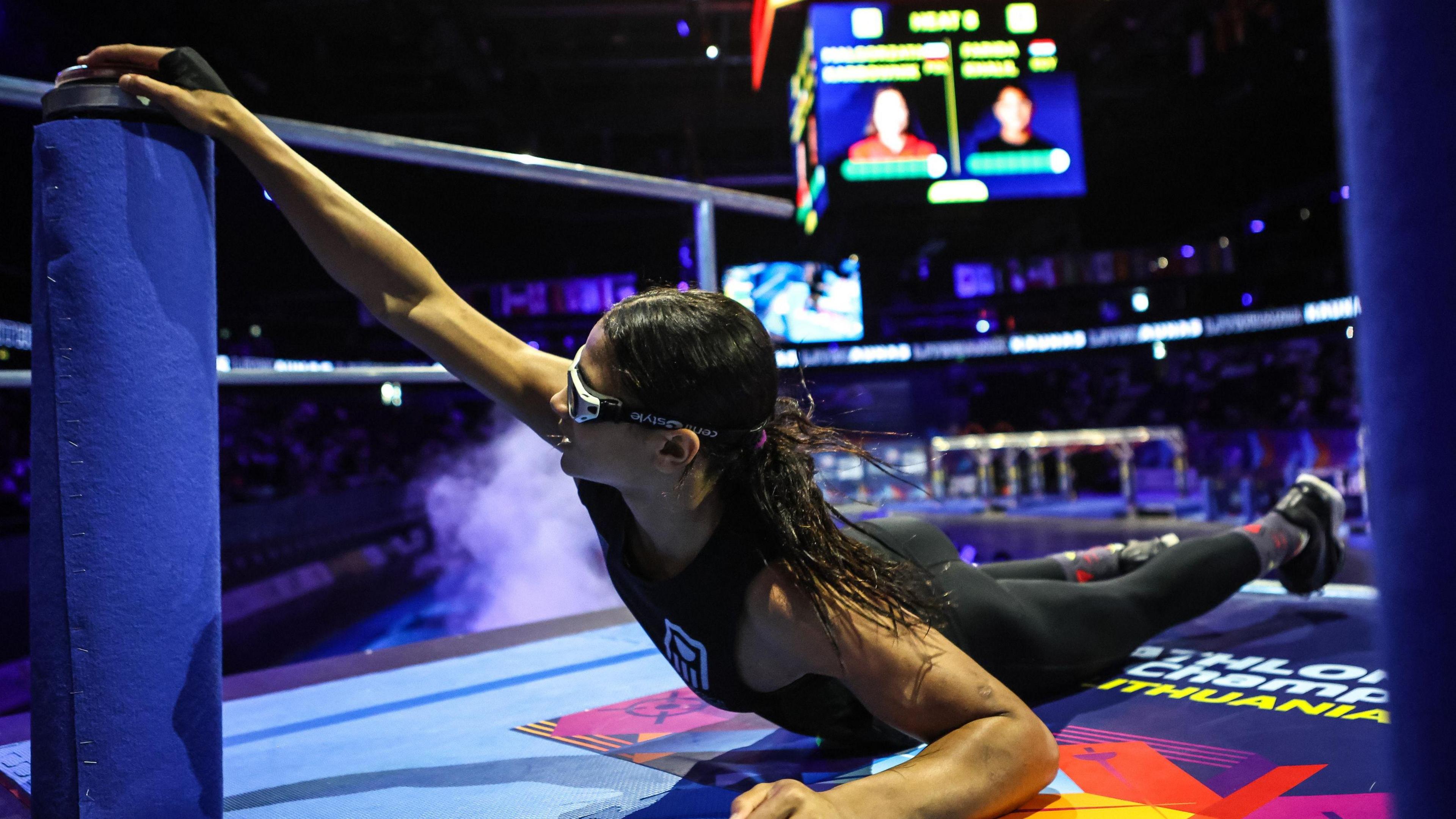 Farida Khalil, wearing sports glasses, a black vest and black leggings, is seen diving close to the ground as she reaches up and attempts to push a buzzer on top of a blue plinth with her right hand. Out of focus in the background a large digital scoreboard and some elements of an obstacle race can be seen inside an indoor arena