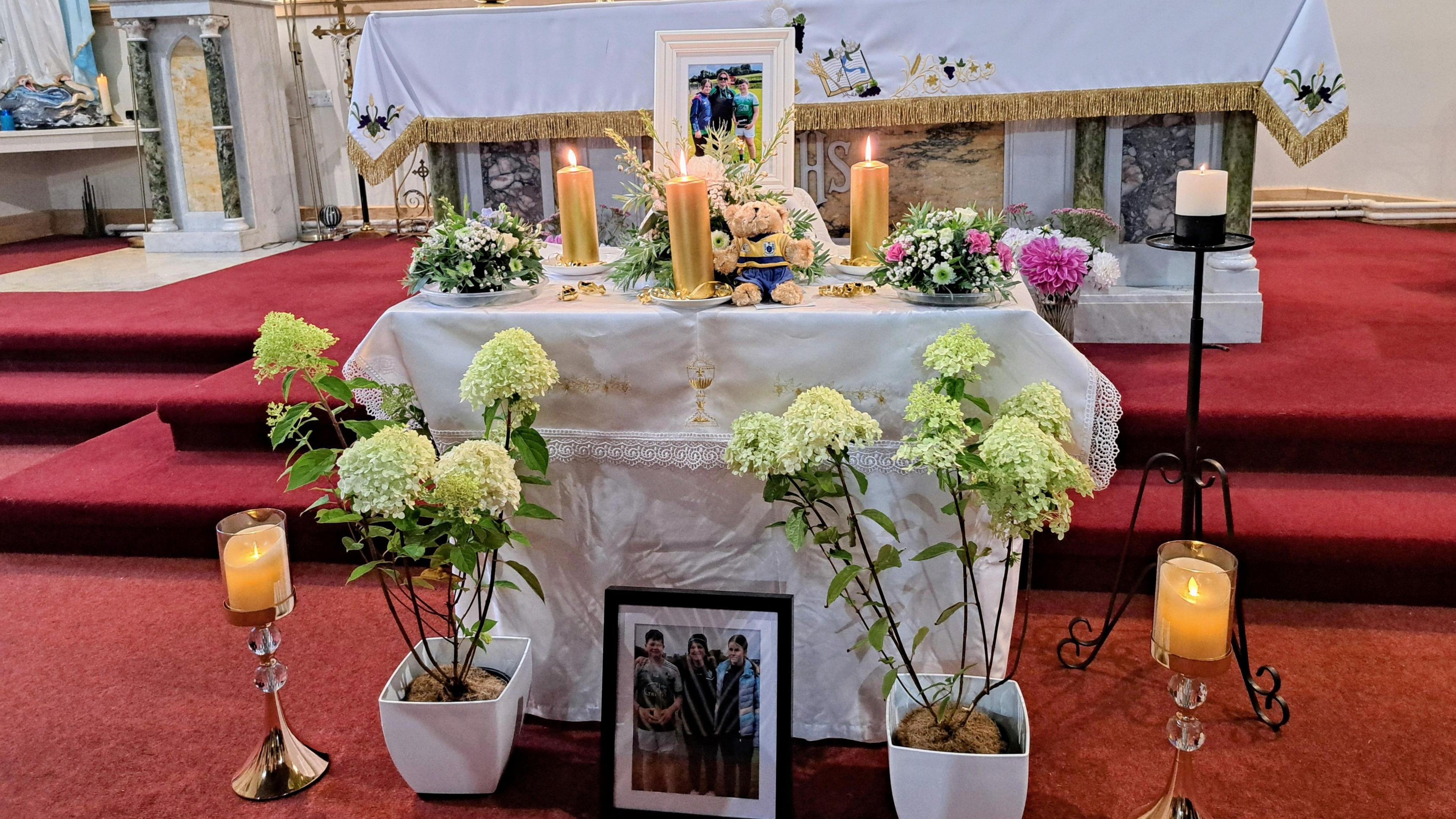 An altar in a church. with three candles lit on top, alongside a teddy and a photo of a mother and two teenagers. Flowers are sitting around the altar.