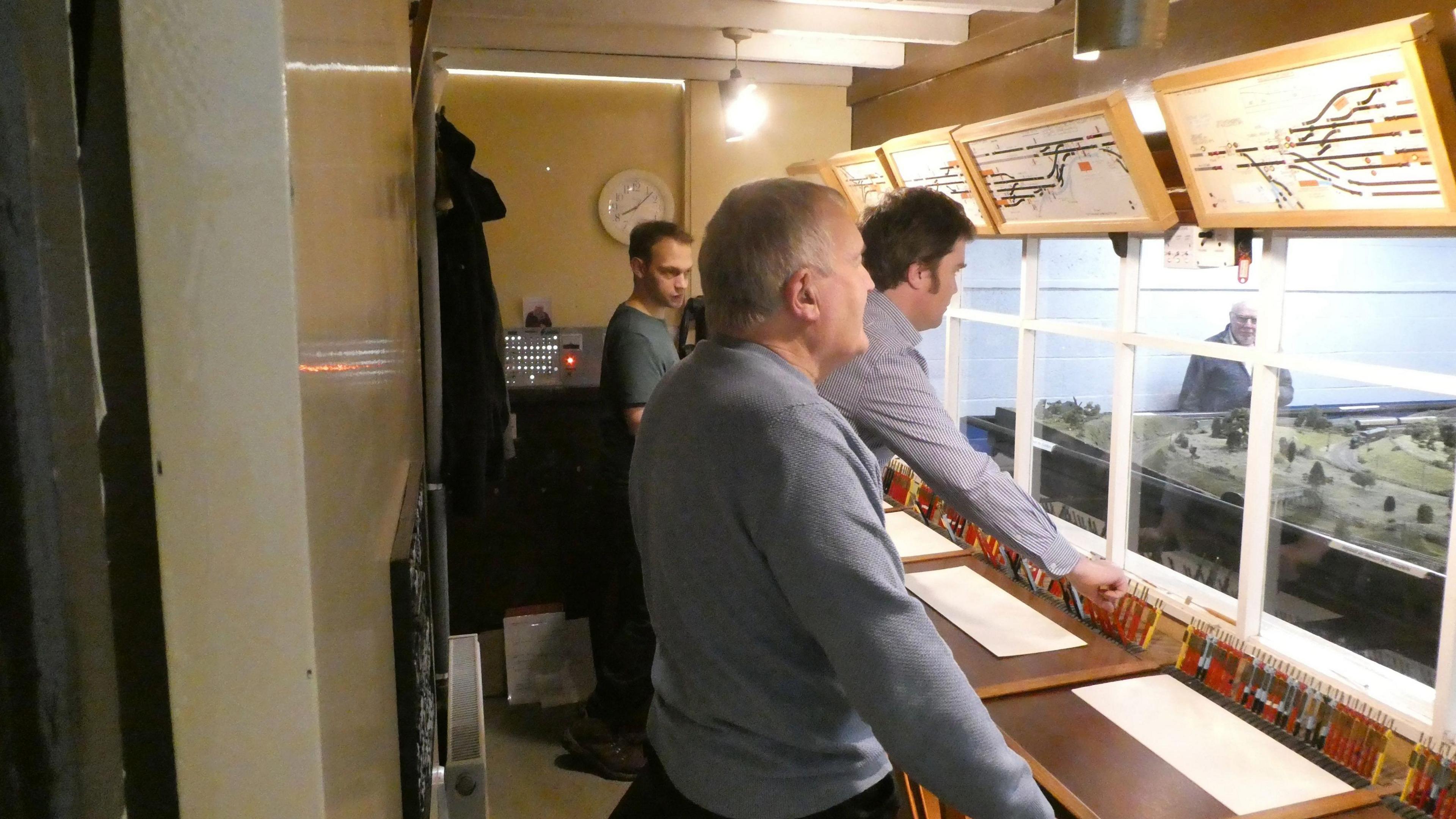 Three men stand inside the signal box. They are controlling lever signals and observing layout signals on the wall.
