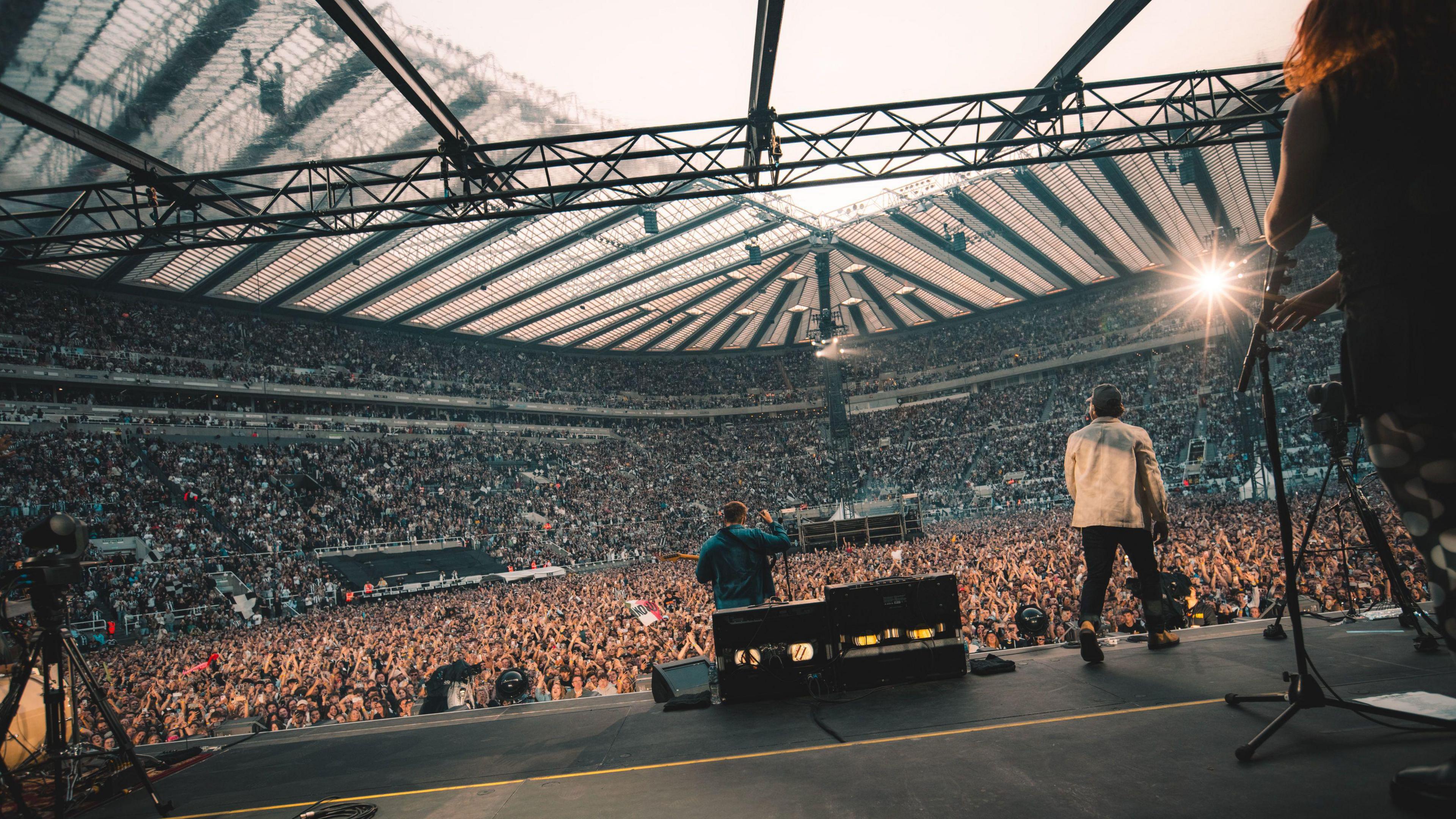 Sam Fender with his back to the camera on a stage singing into the microphone and holding a guitar. A huge crowd of people can be seen in front of him. A man is to his right on stage.