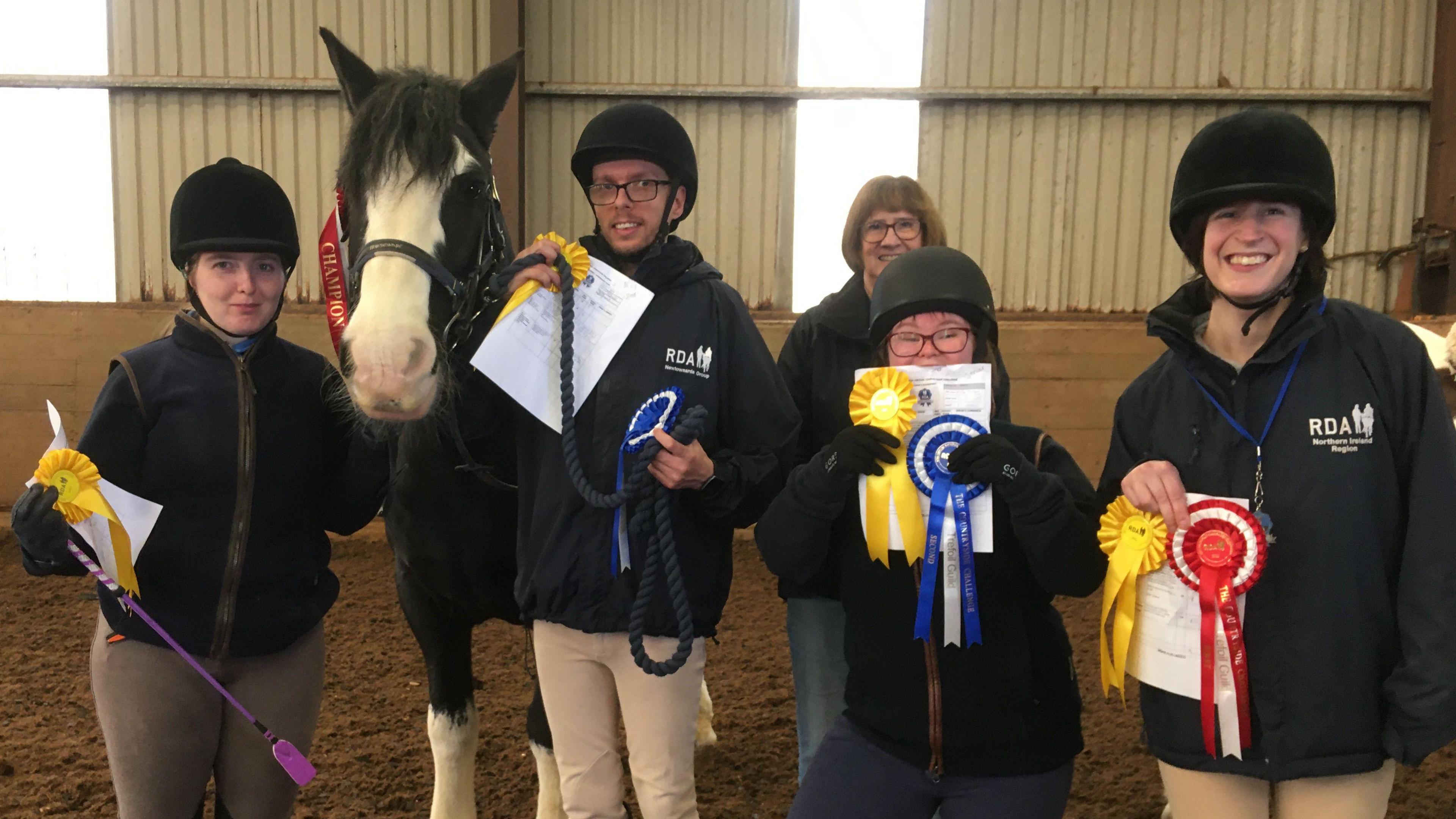 Four horse riders smiling at the camera. There is also a women in the background. The four riders are wearing navy tops RDA branded tops and are holding coloured rosettes. Charlie the horse is standing to the left of one of the riders and is wearing a red champion ribbon on his head.