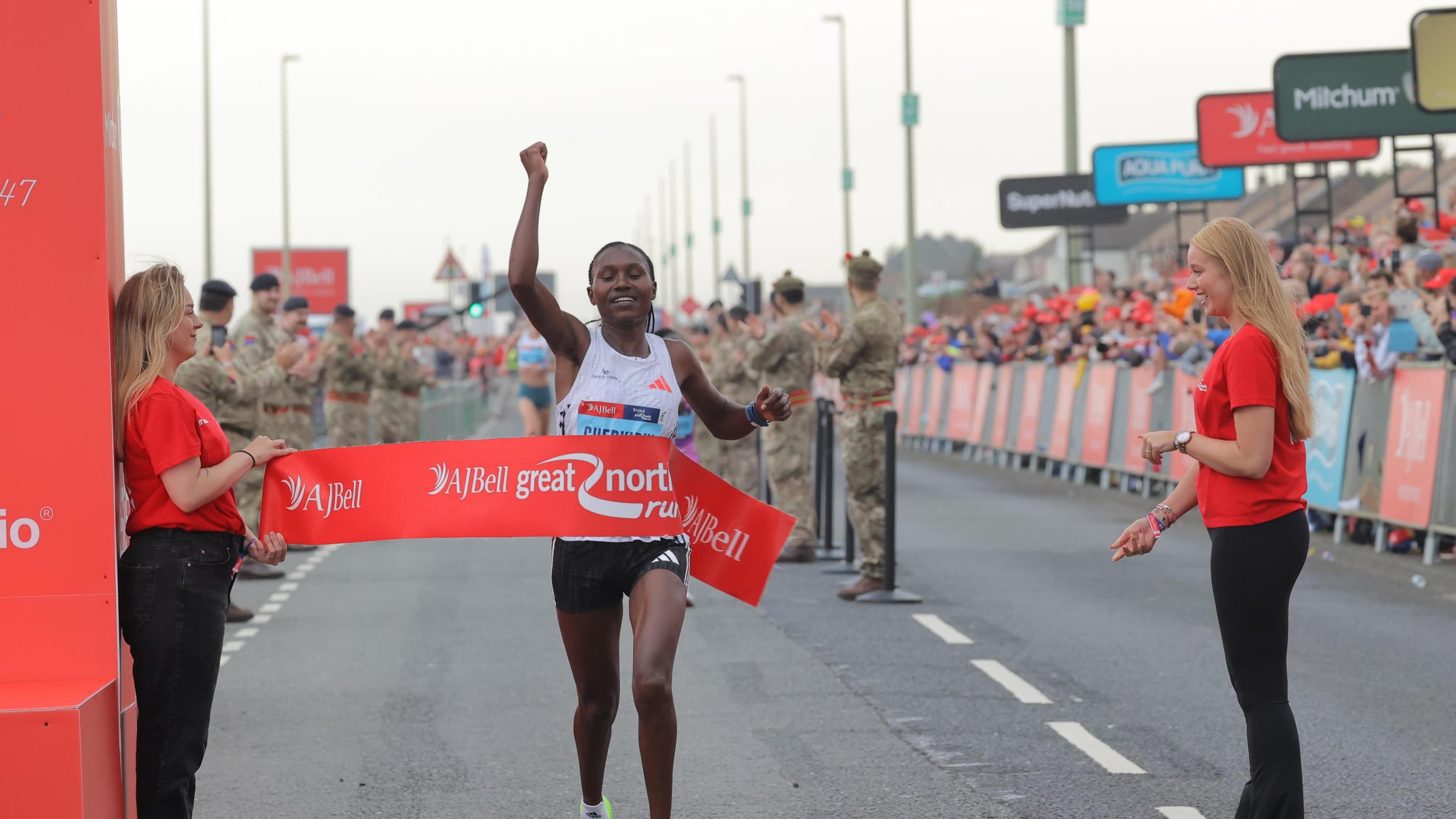 Sheila Chepkirui in runner's clothes crossing winning line with arm in the air in joy. Red finishing line banner wraps over her torso. Crowds cheering in background.