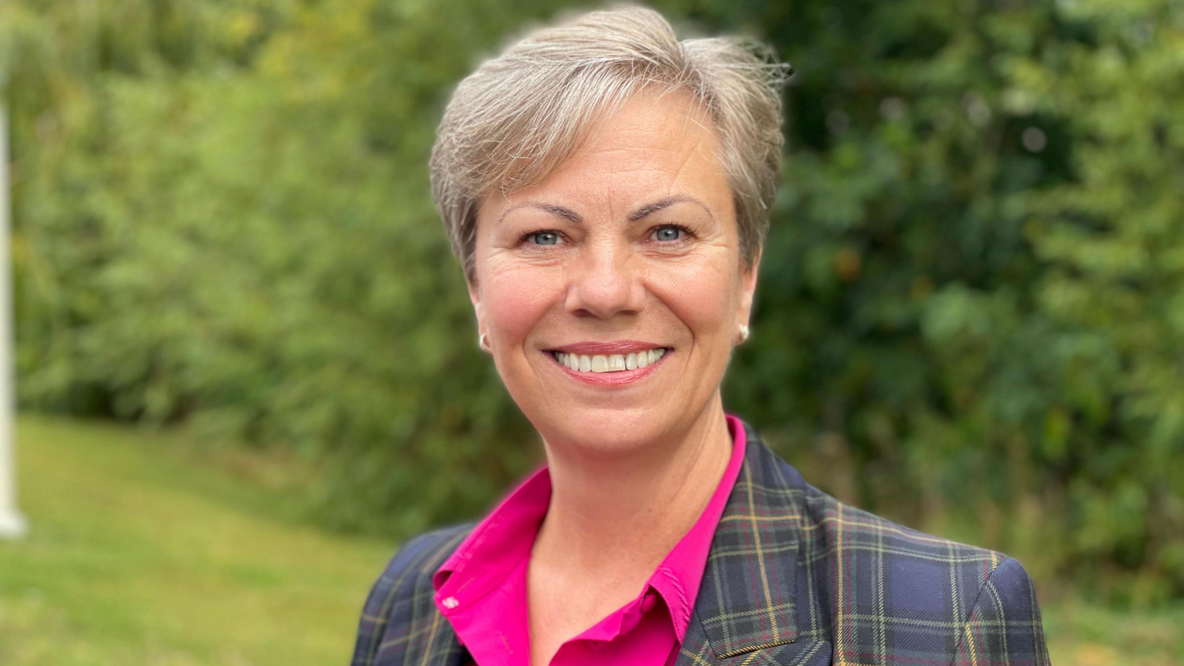 A woman with cropped-grey hair, a pink blouse and a tartan blazer, standing on a lawn in front of a row of tall bushes